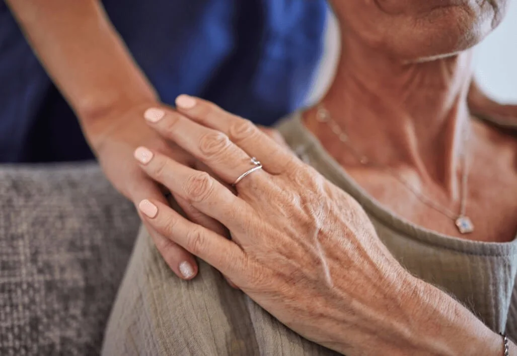 A woman with short grey hair wearing a grey top, showing her hand with a silver ring on her ring finger. Someone is gently touching her shoulder from behind.