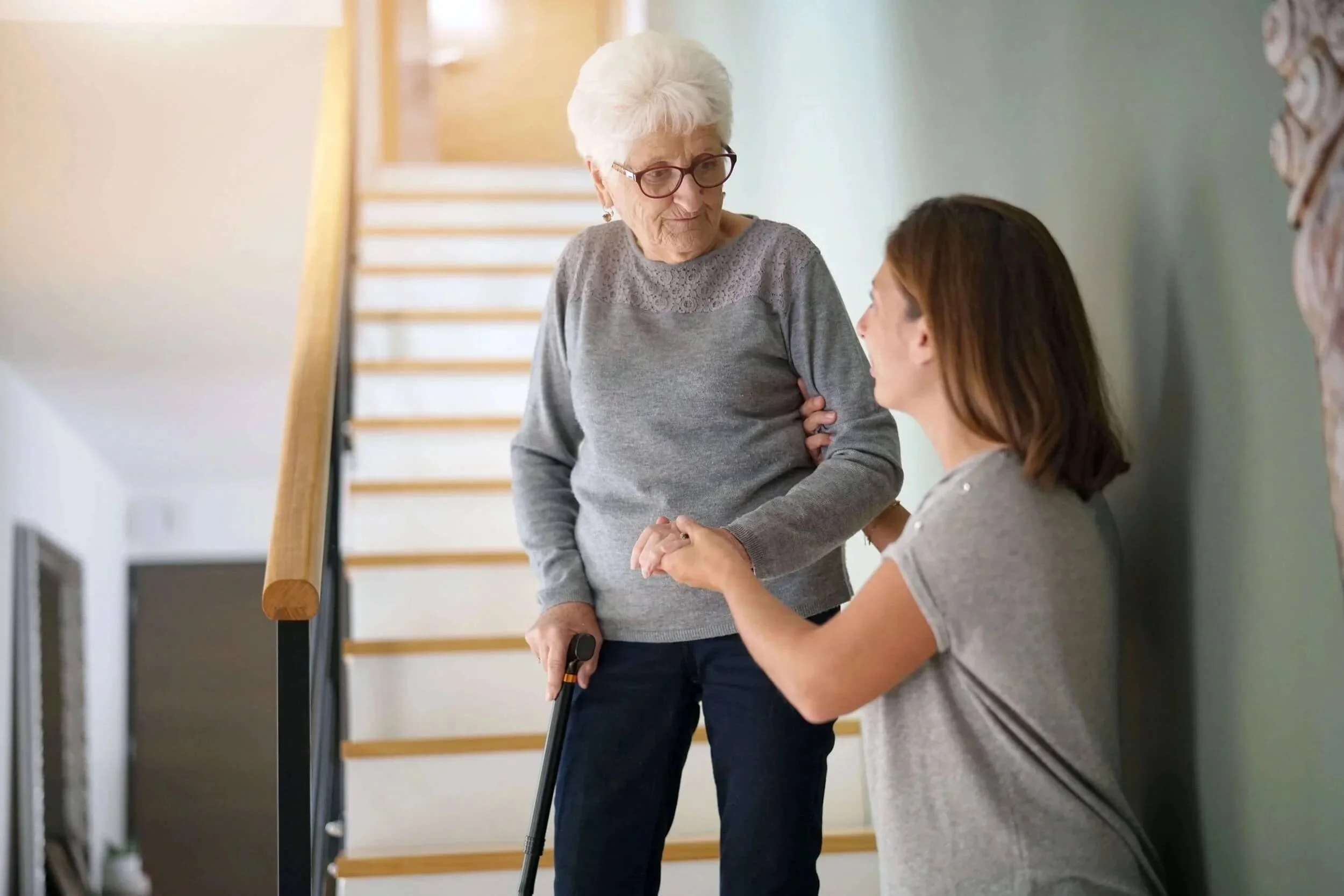 An elderly woman with white hair and glasses helping a younger woman with brown hair who is sitting on the floor with a walking cane in a hallway with a staircase in the background.