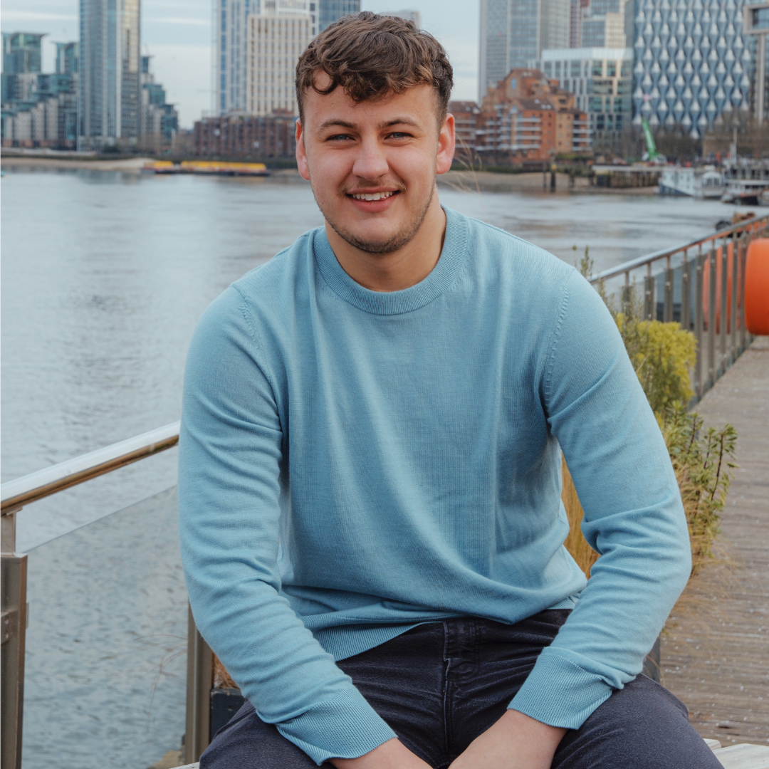 A young man sitting on a bench near a river with a city skyline in the background, smiling at the camera.