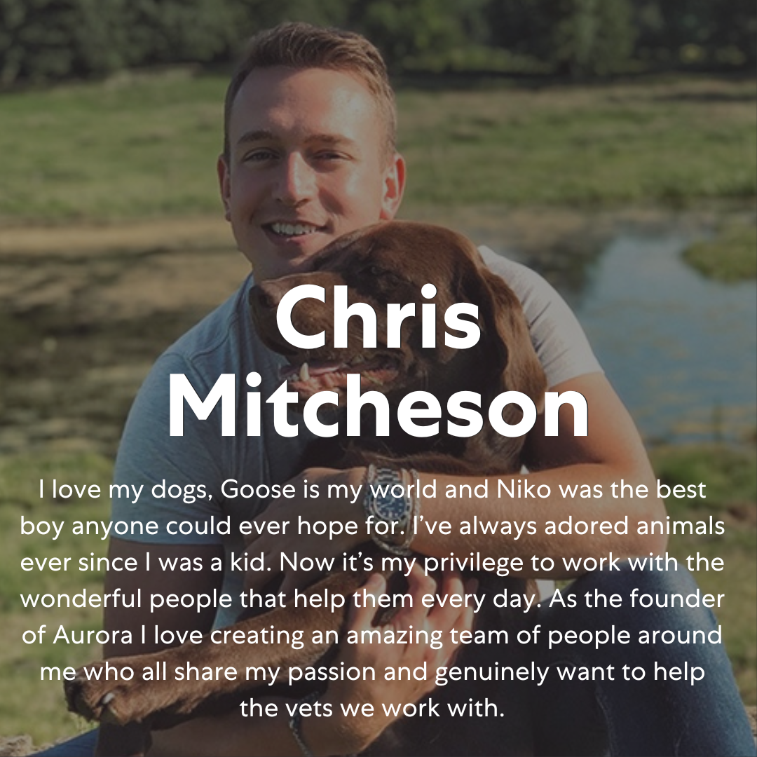A man with short hair smiling outdoors, holding a brown dog with floppy ears in his arms, with a pond and greenery in the background.
