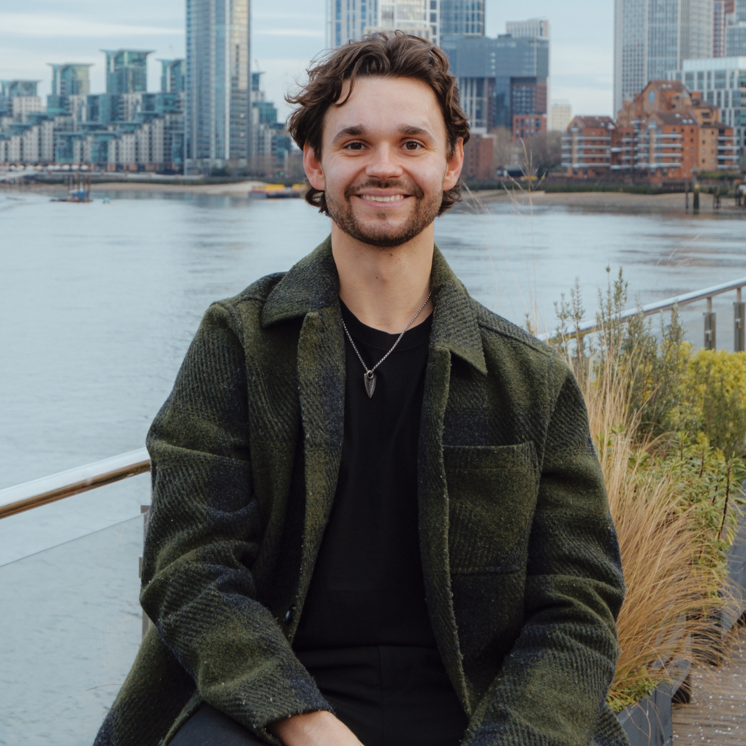 Young man with dark curly hair and a beard smiling, wearing a green plaid jacket and a black shirt, sitting outdoors near a river with city buildings in the background.