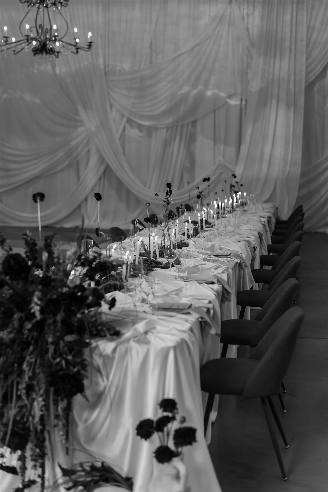 Long banquet table decorated with flowers, candles, and tableware, set for an elegant event in a draped curtain venue with a chandelier overhead.