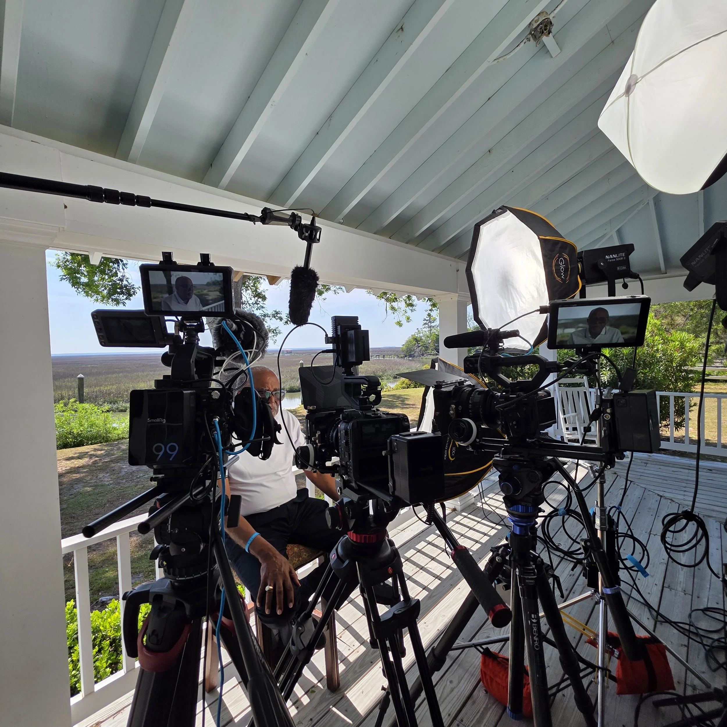 Video production setup with multiple professional cameras, monitors, microphones, and studio lights on a seaside porch