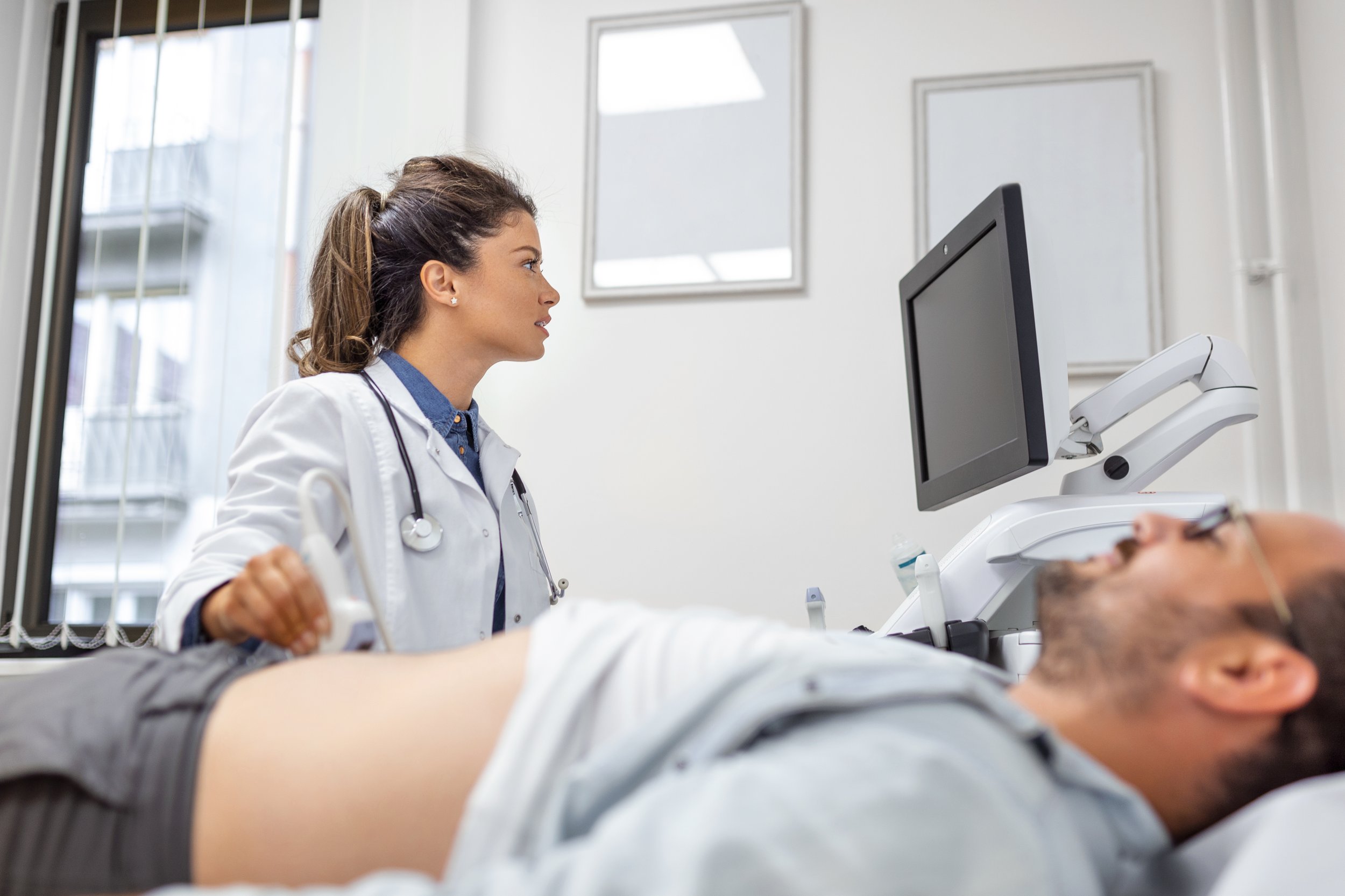 A female healthcare professional performs an ultrasound scan on a male patient lying on a medical bed.
