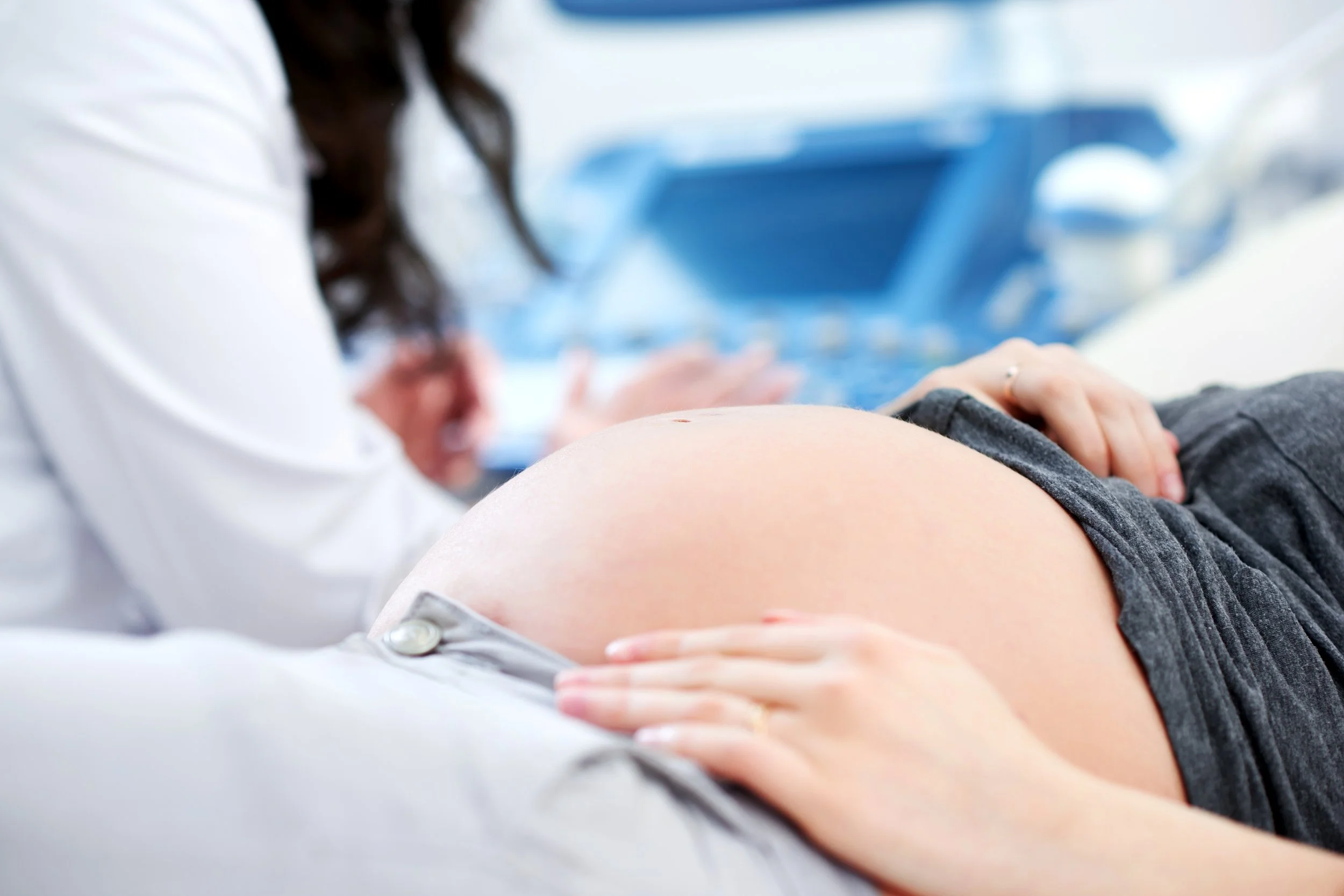 A close-up of a pregnant woman's belly during a prenatal ultrasound exam, with a medical professional's hand gently resting on her abdomen, and a blue ultrasound machine in the background.