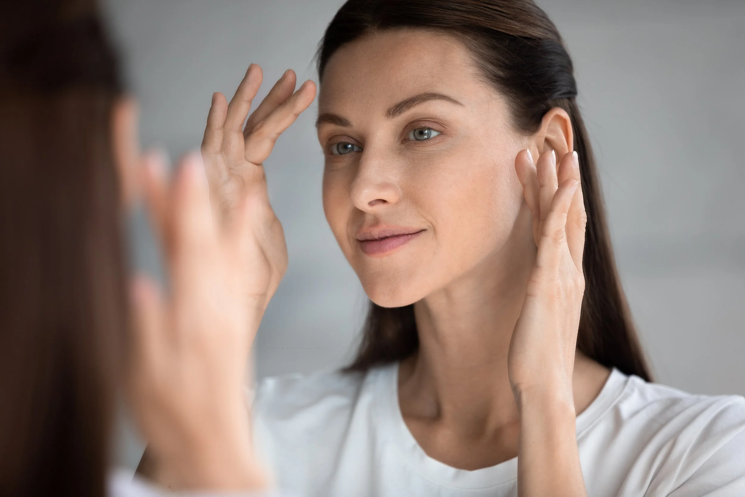 A woman with long dark hair touches her face near her ear and forehead while looking at her reflection.