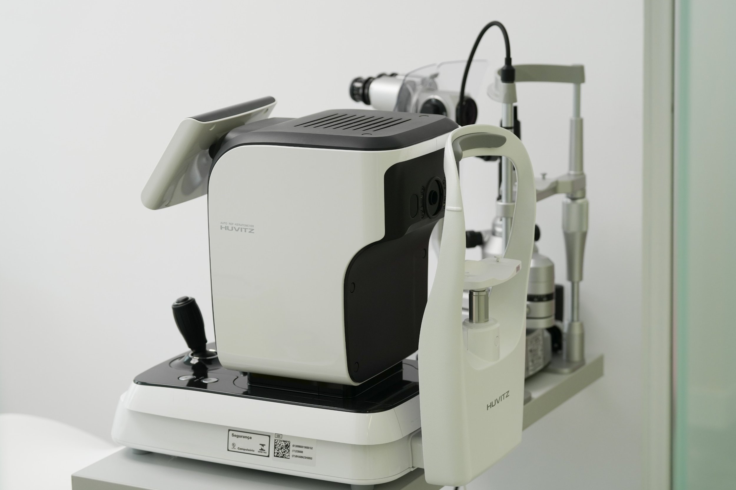An ophthalmic device called a Huvitz autorefractor used for eye examinations, with a joystick, eye rest, and attached equipment, in a medical setting.