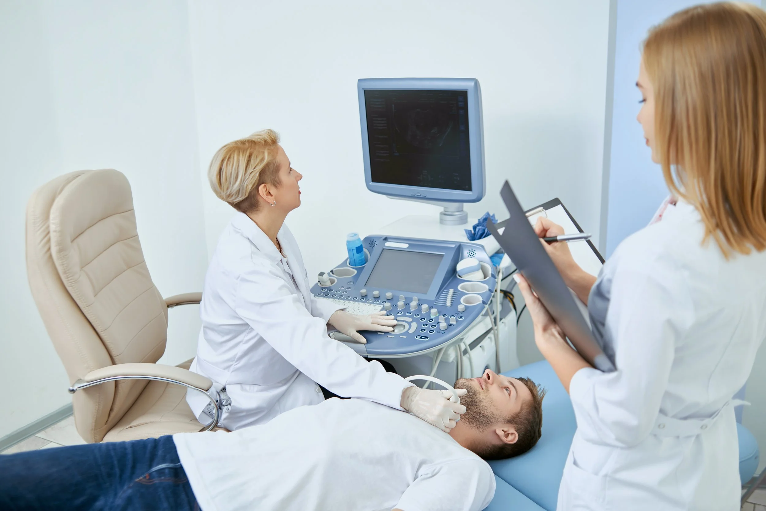 Medical professionals performing an ultrasound examination on a man lying on a hospital bed, with one doctor operating the ultrasound machine and another taking notes.