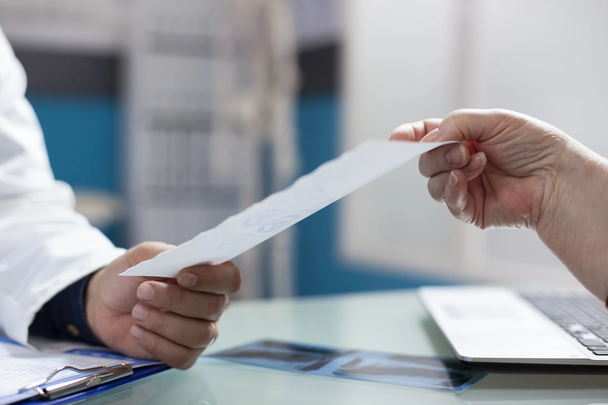 Two people exchanging a piece of paper in an office setting, with a laptop and documents on the desk.