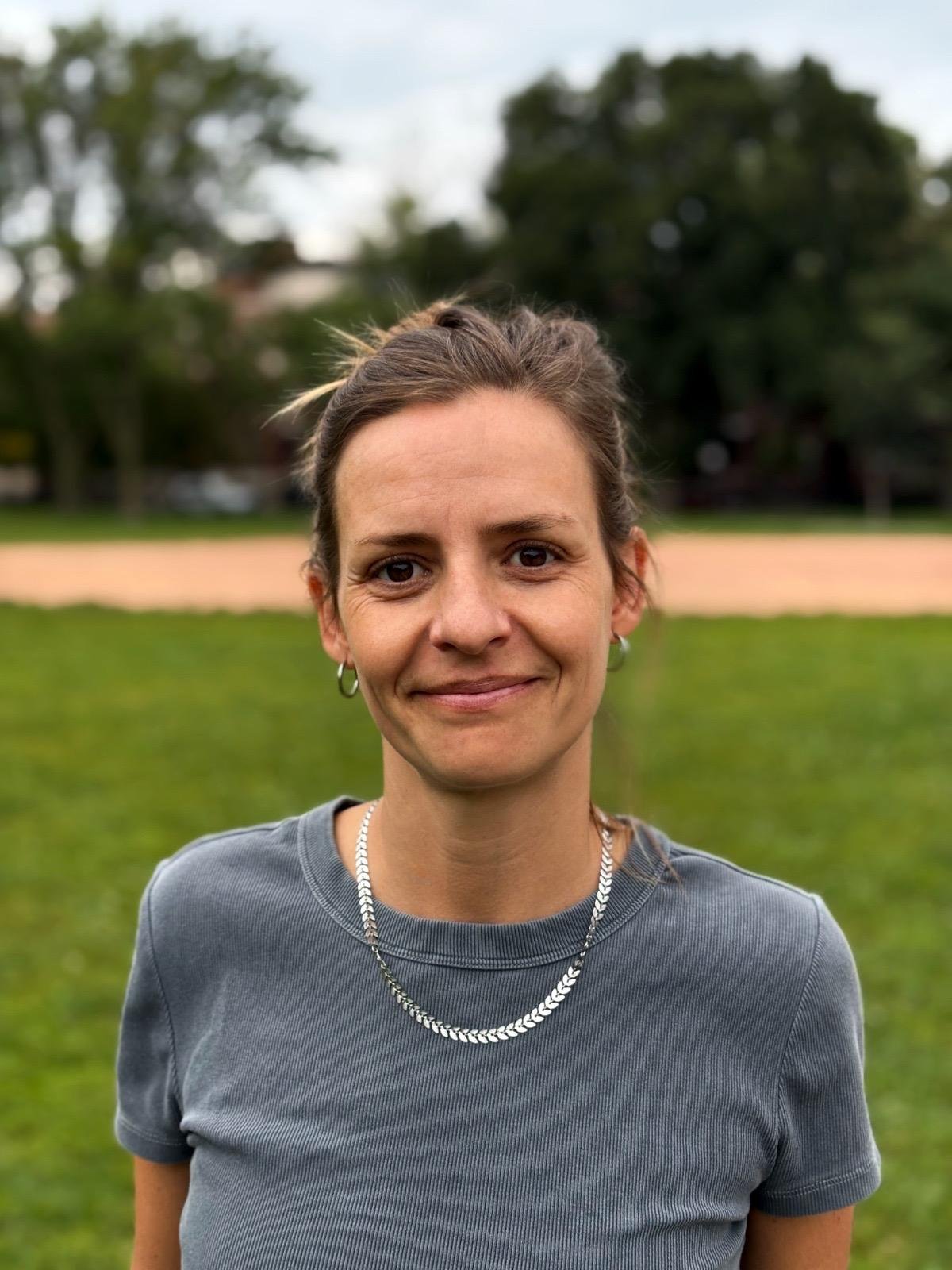 Smiling woman with brown hair tied back, wearing a grey t-shirt, silver necklace, and hoop earrings, standing outdoors on a grassy field with trees and a path in the background.