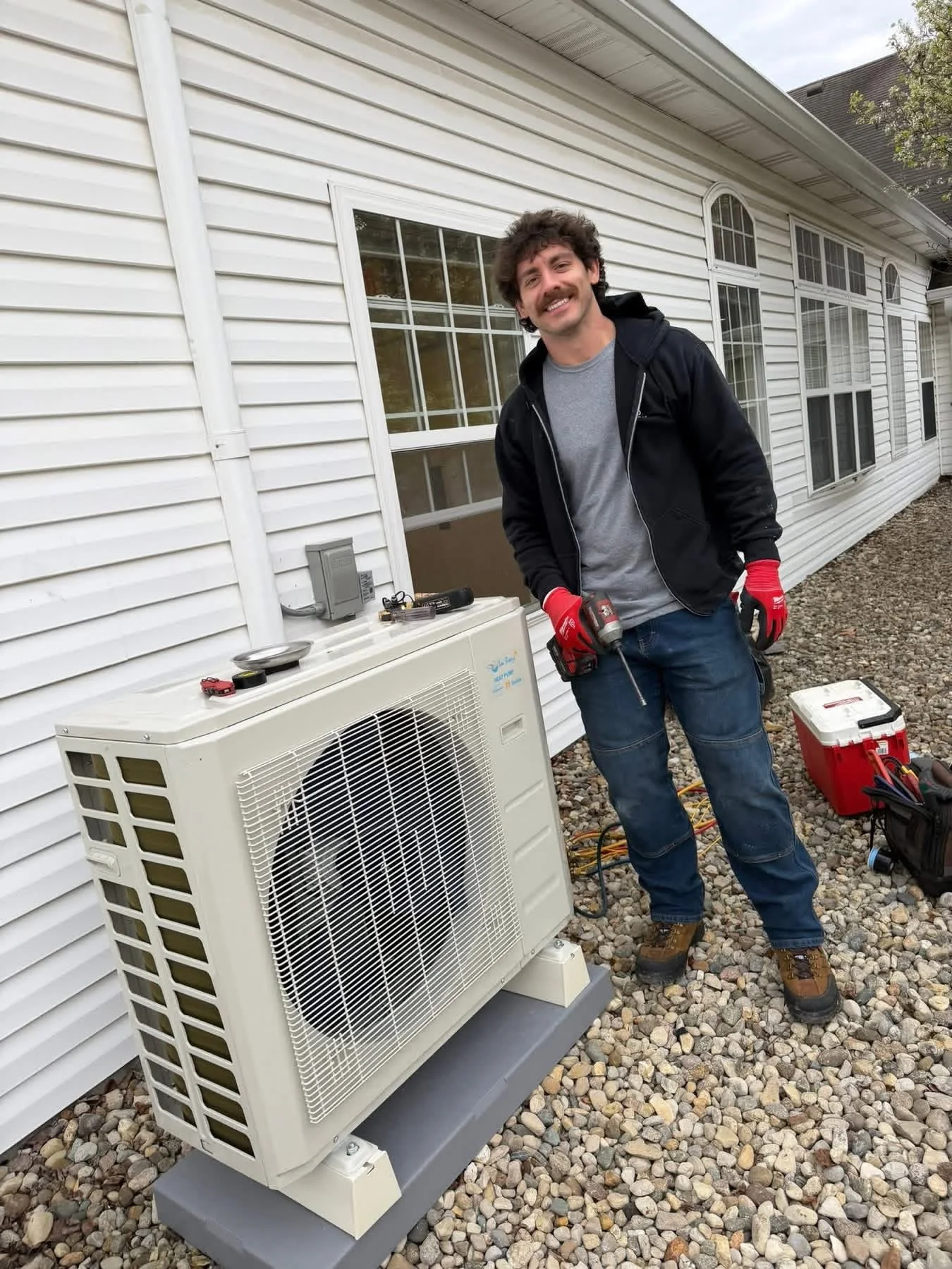 A smiling man in a gray shirt, black jacket, red gloves, and work boots is standing outside a house next to an air conditioning unit. He is holding a tool and appears to be working on the unit. There is a toolbox and other tools on the ground nearby.