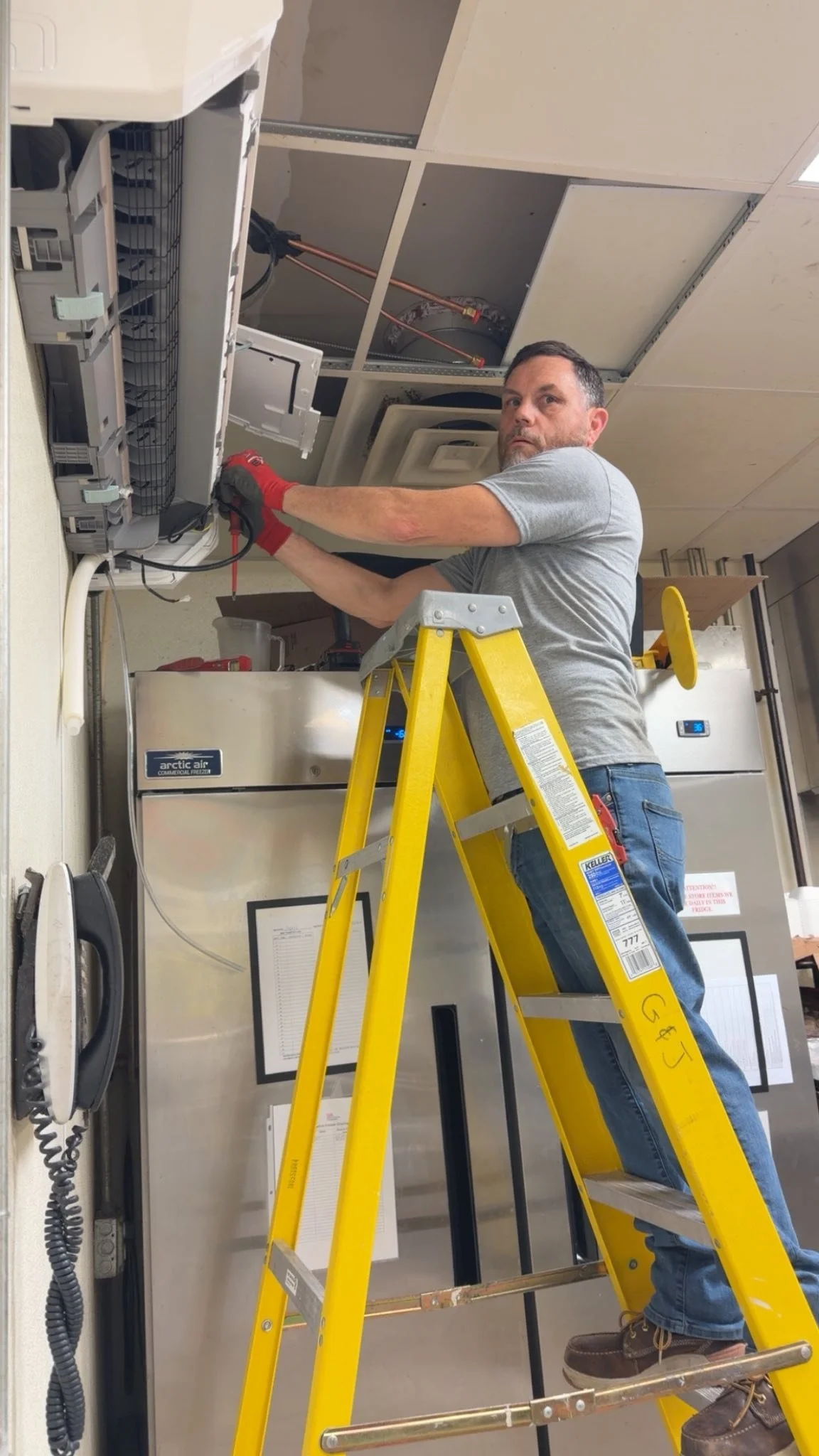 A man on a yellow ladder working on electrical wiring inside a ceiling, wearing a gray t-shirt and jeans.