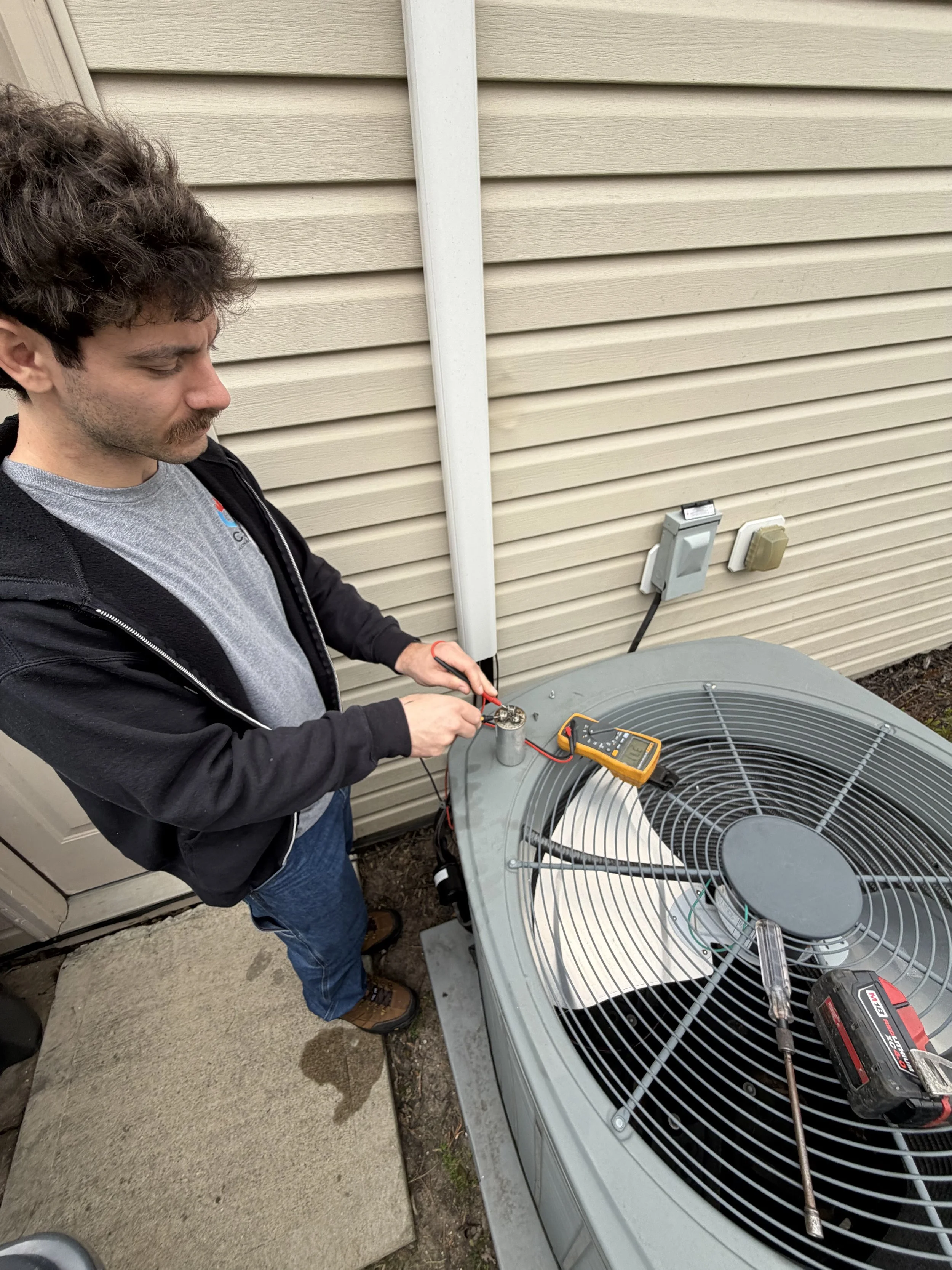 A man performing maintenance or troubleshooting on an outdoor air conditioning unit using a multimeter.