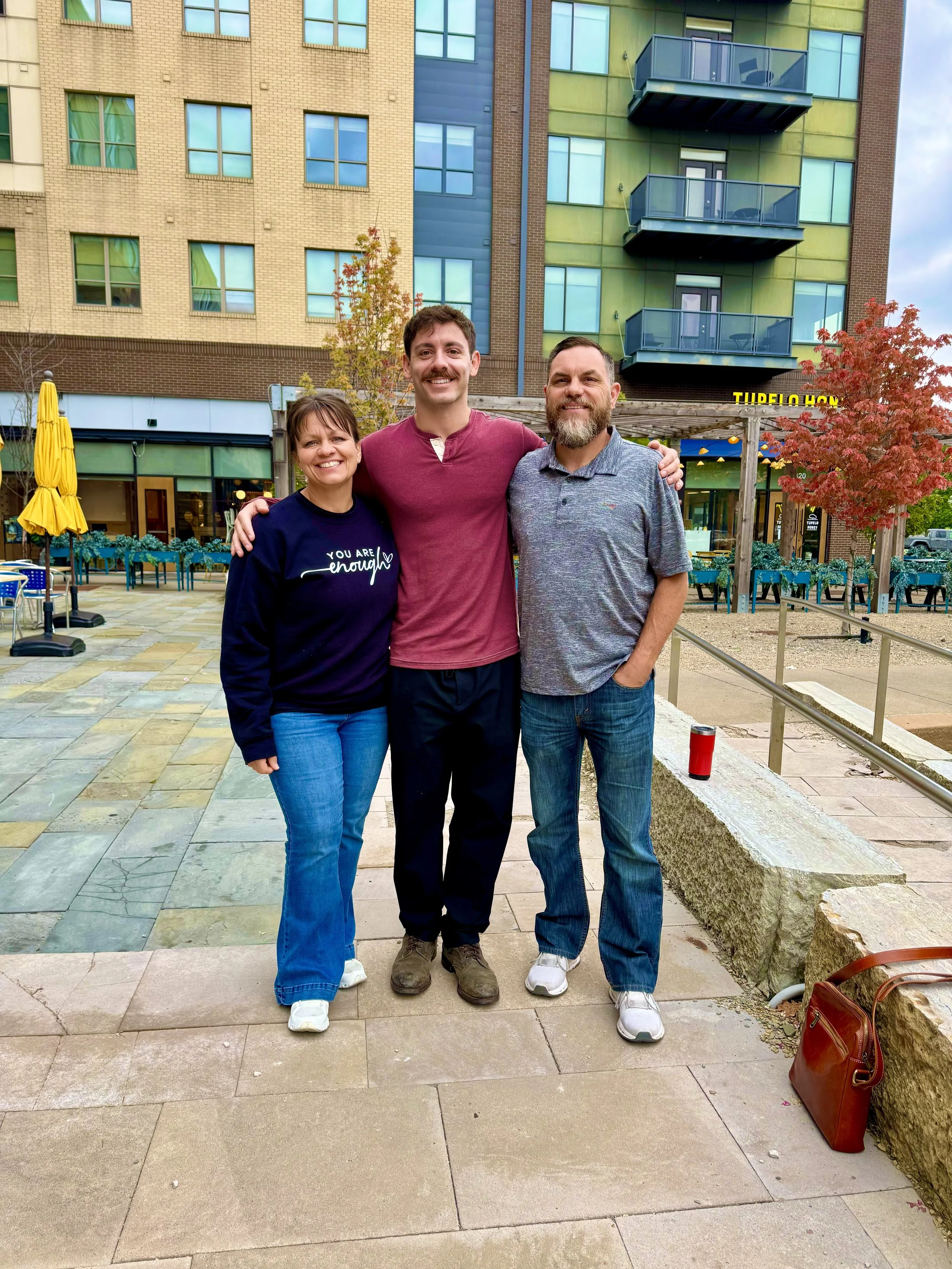 Three people standing outside in an urban setting with colorful buildings in the background, smiling and posing for a photo.