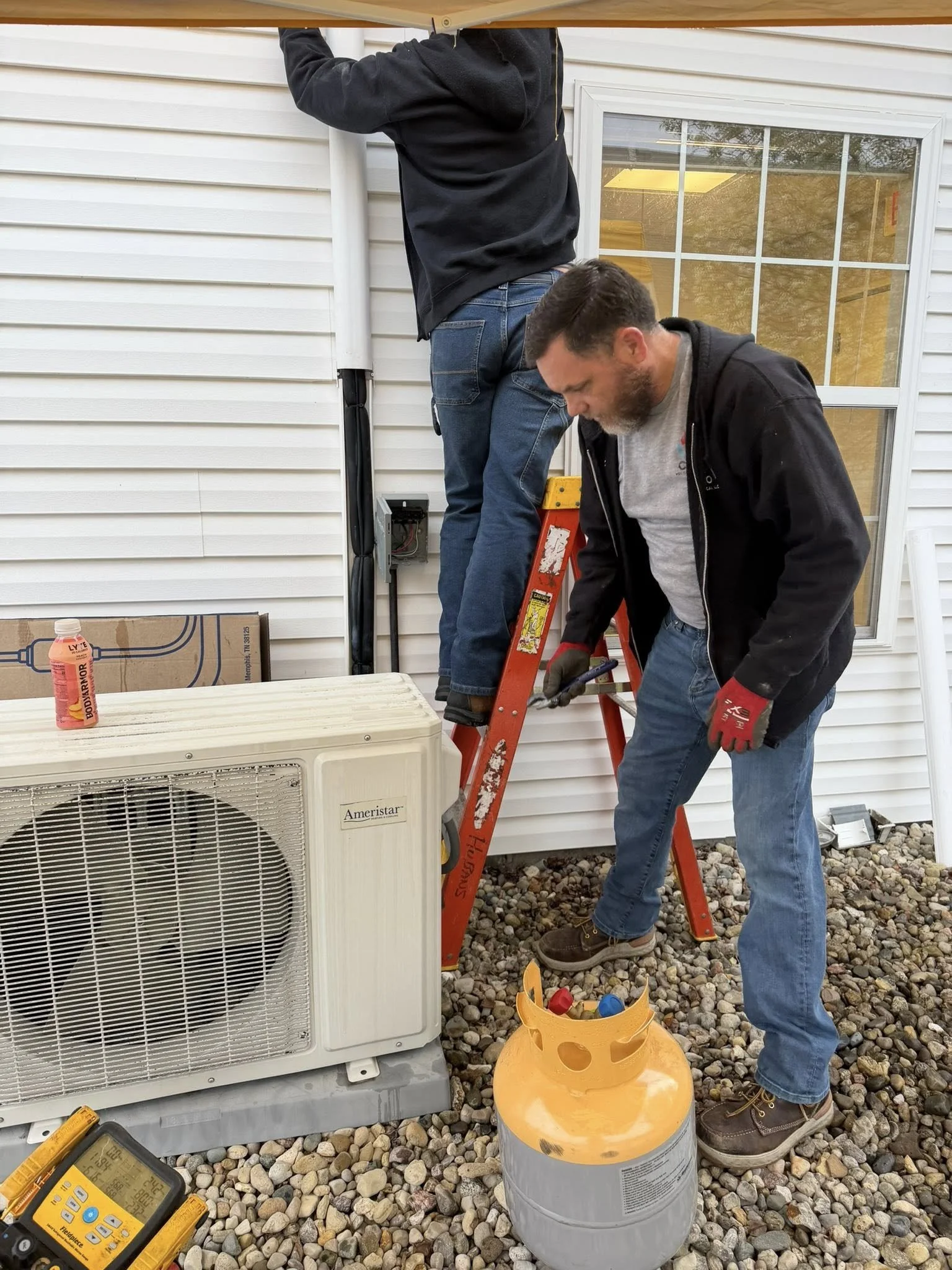 Two men working on an air conditioning unit outside a house. One man is standing on a ladder, and the other is holding a tool near the unit. A can of refrigerant and a digital thermometer are also visible.
