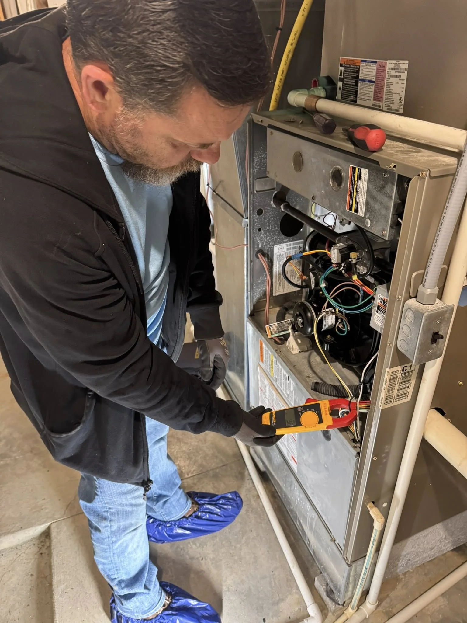A technician in a black jacket, light blue shirt, and blue shoe covers is using a multimeter to test the electrical components inside an open furnace.