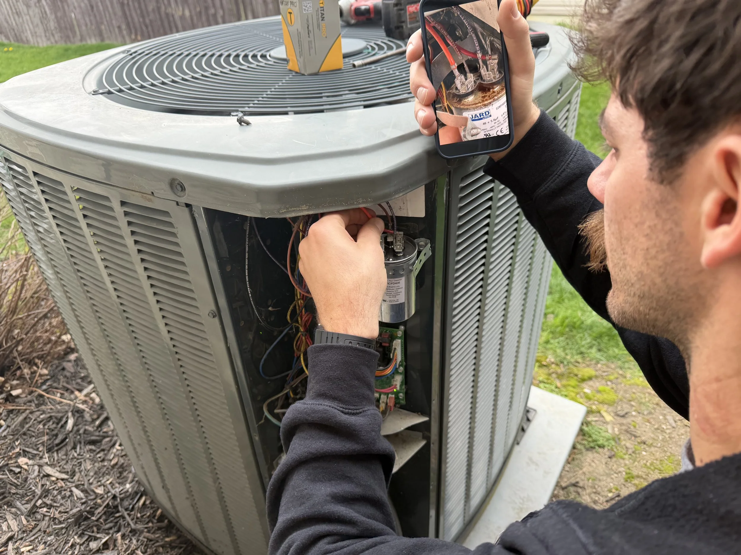 A technician repairing an outdoor air conditioning unit, inspecting the interior components with a screwdriver and taking a photo of the inner wiring with a smartphone.