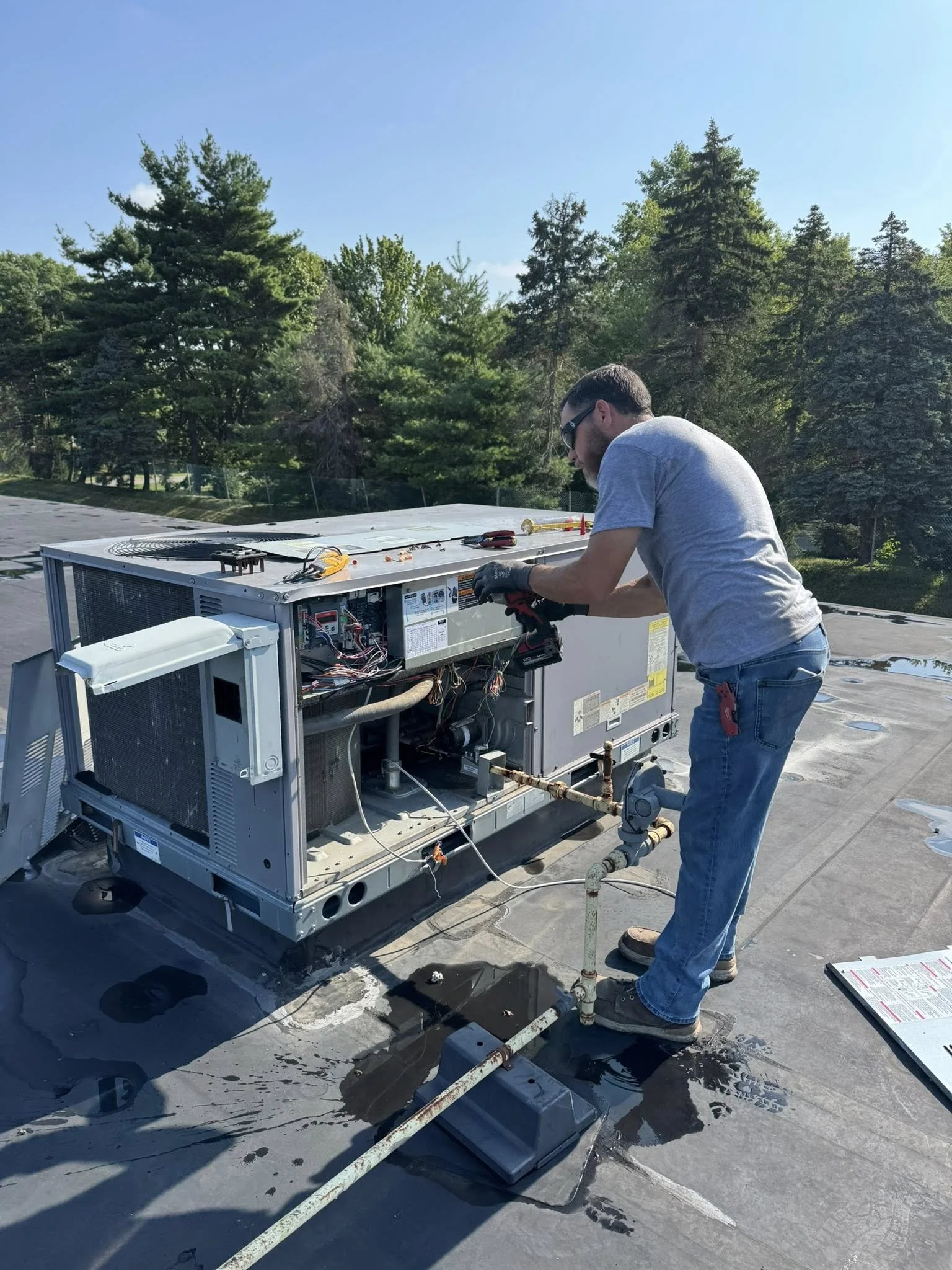 A technician wearing a gray shirt and blue jeans is repairing an air conditioning unit on a flat rooftop, with tools and equipment around him and trees in the background.
