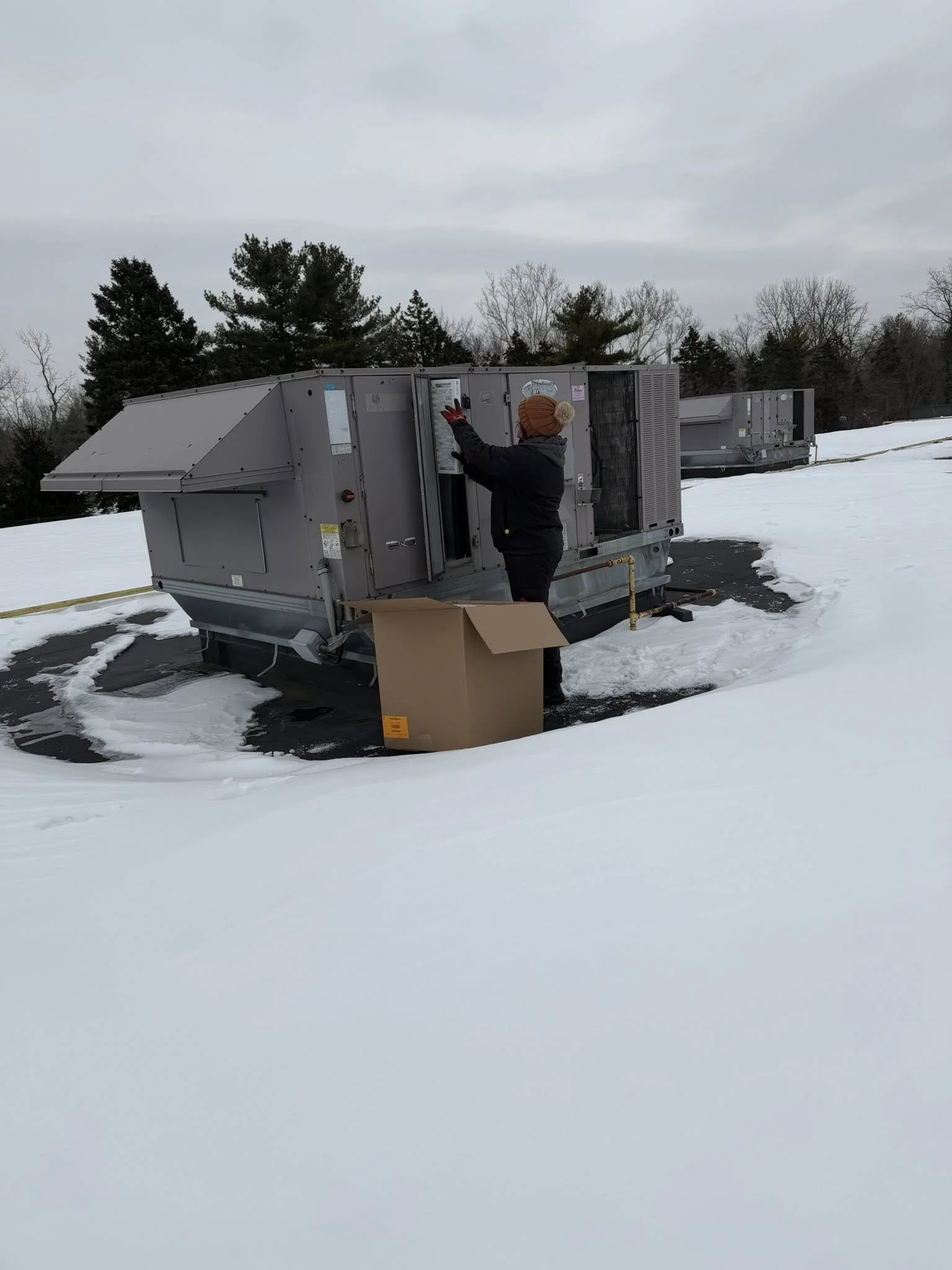 A person in winter clothing and a knit hat working on an HVAC unit outdoors in a snowy landscape, with an open cardboard box nearby.