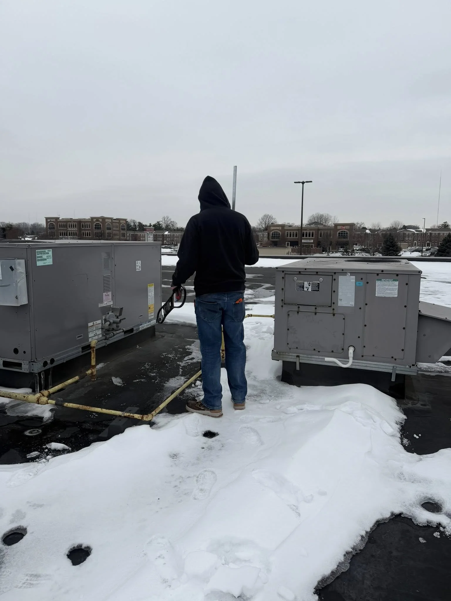 Person wearing a black hoodie and jeans standing on snow-covered rooftop near HVAC units.
