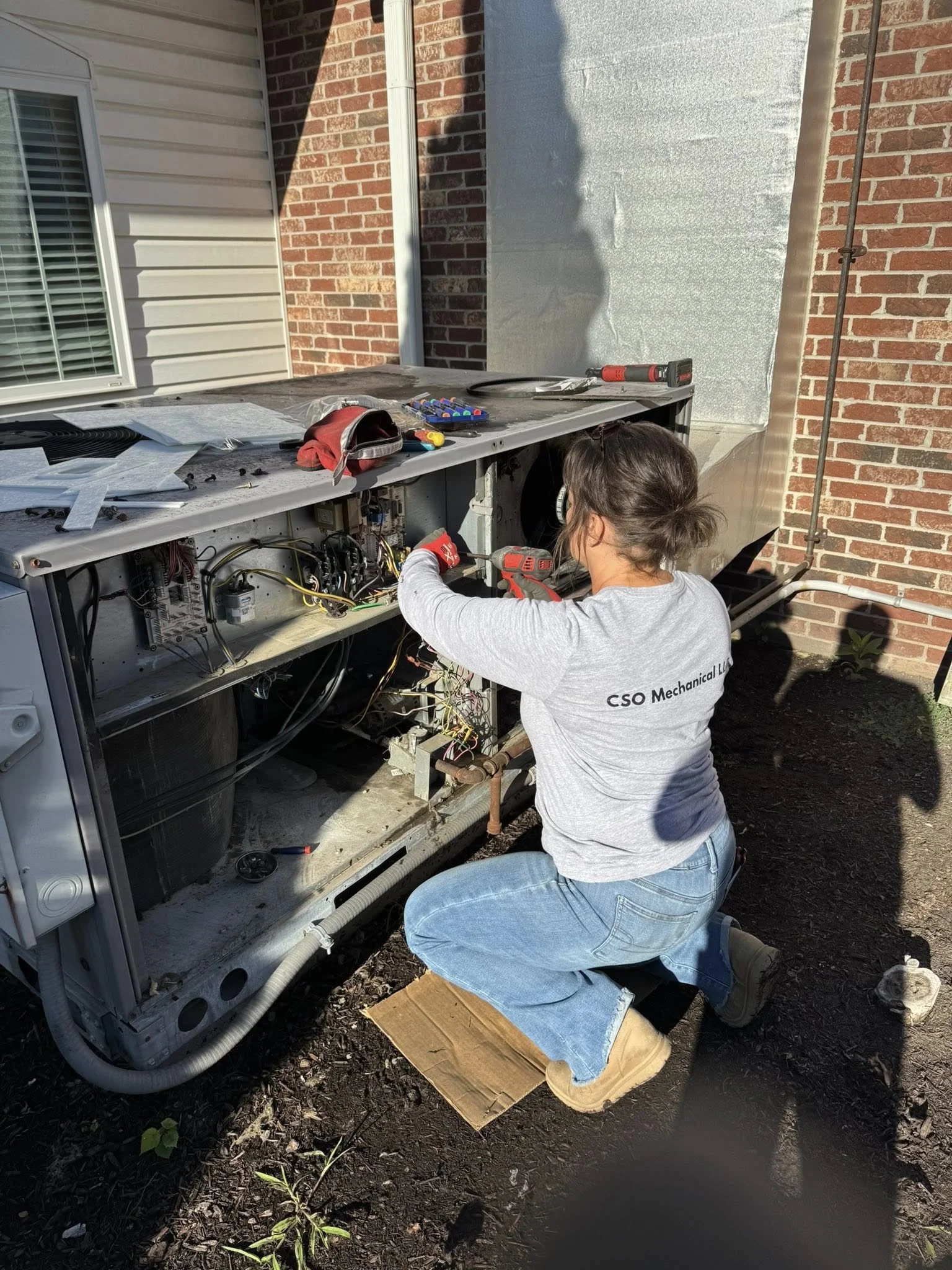 A woman working on an outdoor air conditioning unit, using tools and surrounded by various repair equipment.