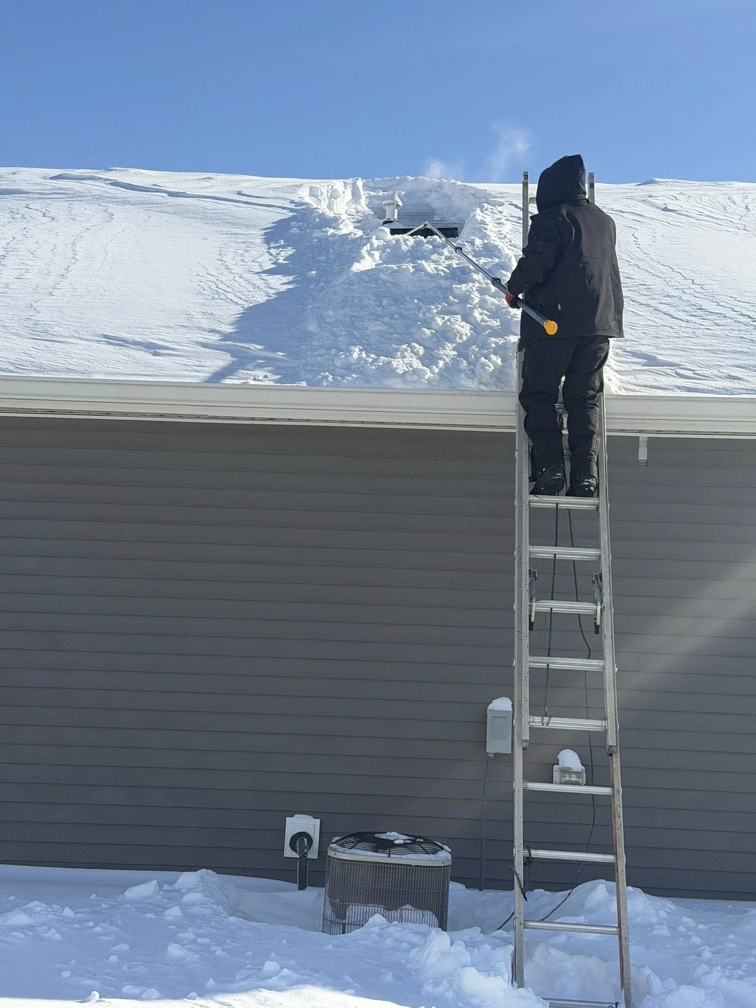 Person wearing a black winter coat and pants, standing on a ladder, clearing snow from the roof of a house with a snow rake in winter.
