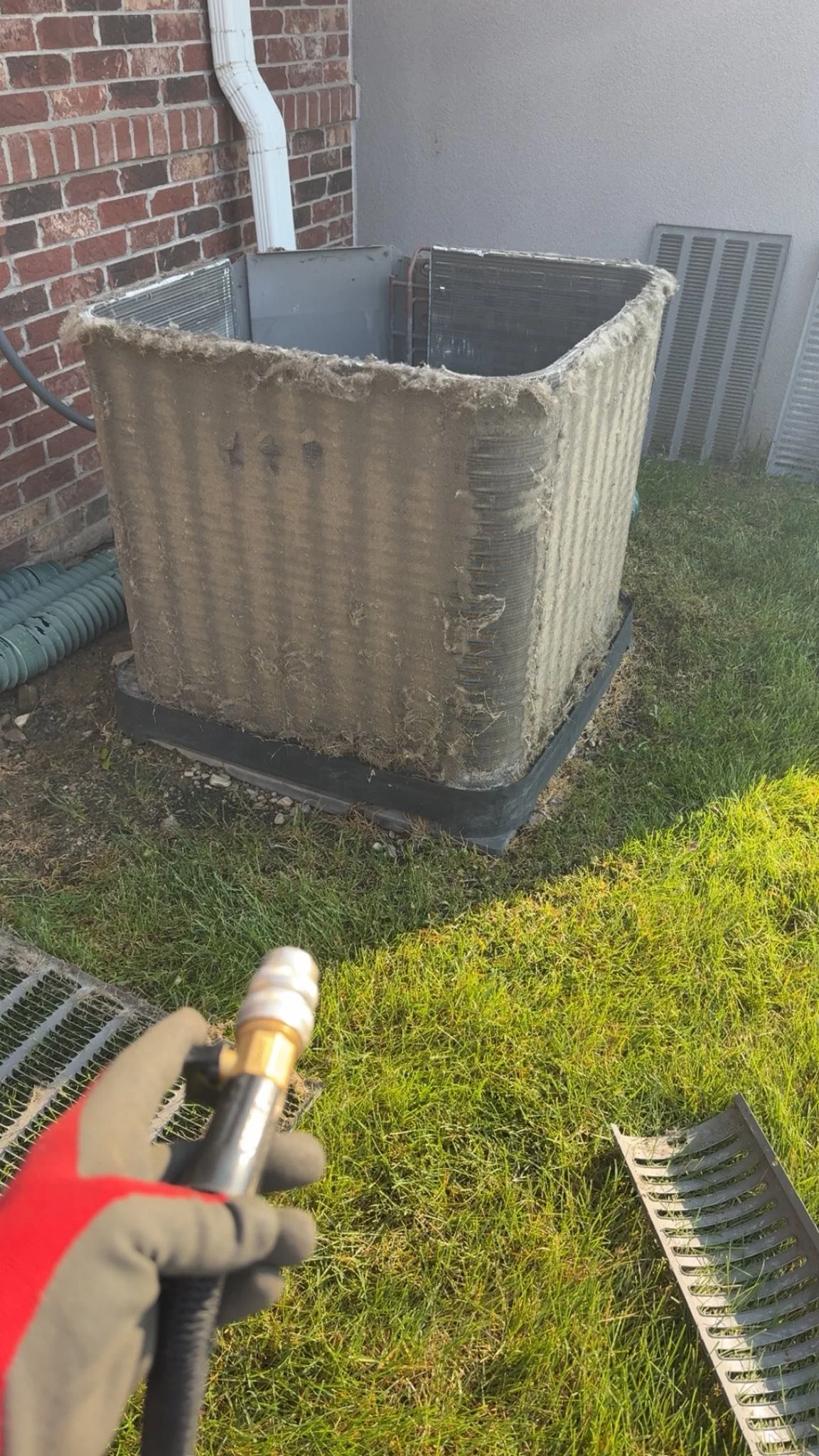 An outdoor air conditioning unit with a coil that is dirty and covered in dust and debris. A person in gloves is holding a tool, possibly for cleaning or repairing the unit. There are grass, a drain grate, and a brick and stucco wall in the background.