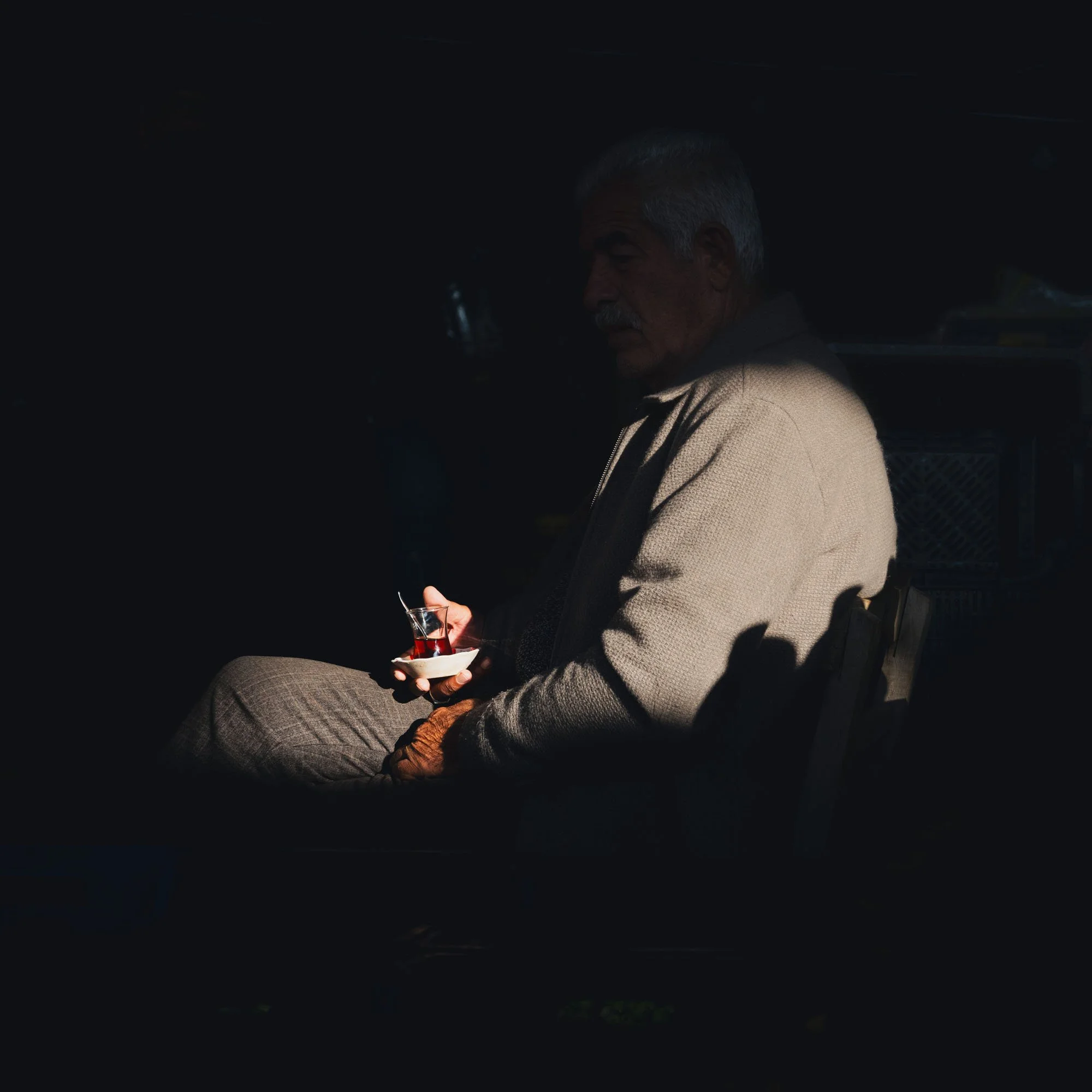 An older man in Istanbul sipping Turkish tea in a sharp beam of light. The heavy chiaroscuro lighting creates visual ambiguity, focusing the viewer's attention on the quiet narrative of the main event.