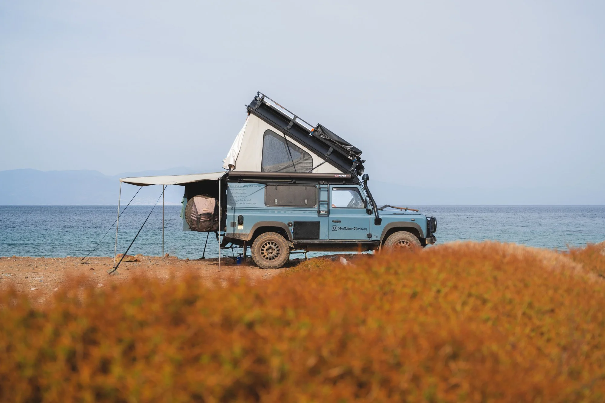 Side profile of a Land Rover Defender with a rooftop tent deployed at a coastal campsite in Türkiye. The out-of-focus orange foreground creates a visual frame, utilizing depth of field to focus on the vehicle.