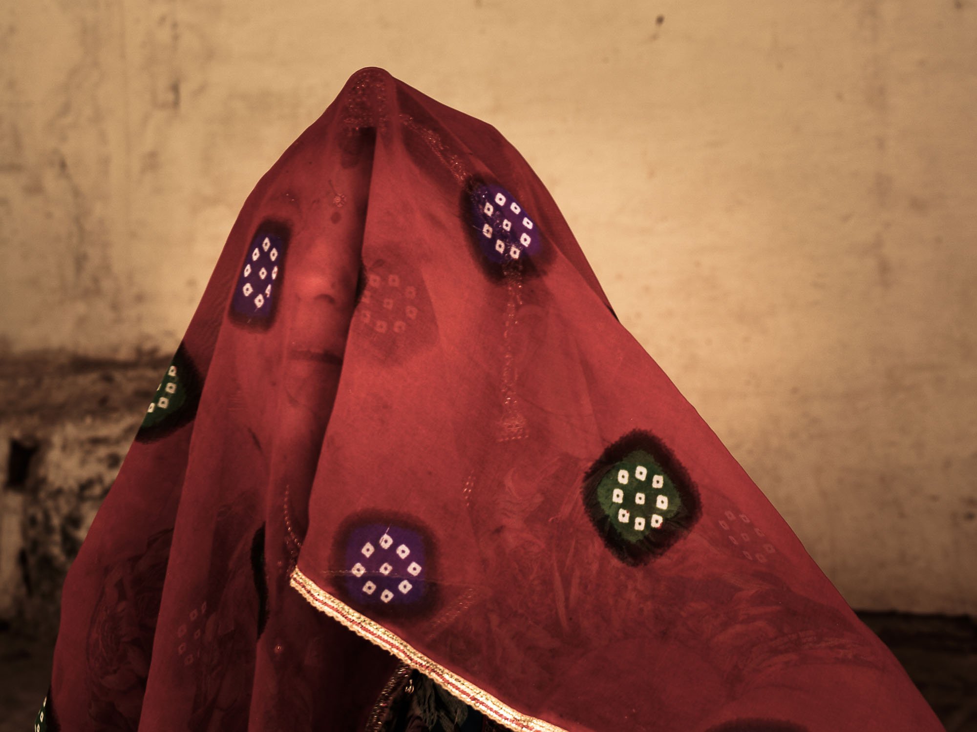 tight portrait of a woman in Rajasthan partially obscured by a sheer crimson veil. This frame demonstrates the 'Curiosity Gap' by withholding the subject's identity while providing enough of a human signal to trigger the seeking system