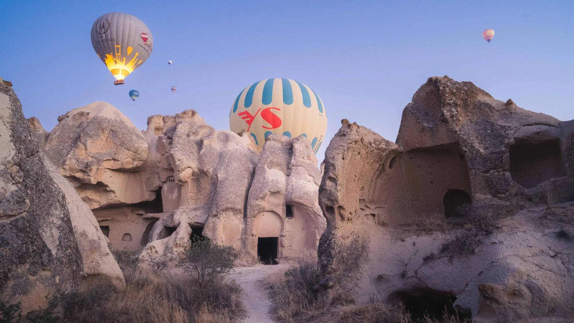 Vibrant hot air balloons looming over ancient rock-cut cave dwellings and fairy chimneys in Cappadocia, Türkiye, highlighting the tension between the ephemeral and the ancient