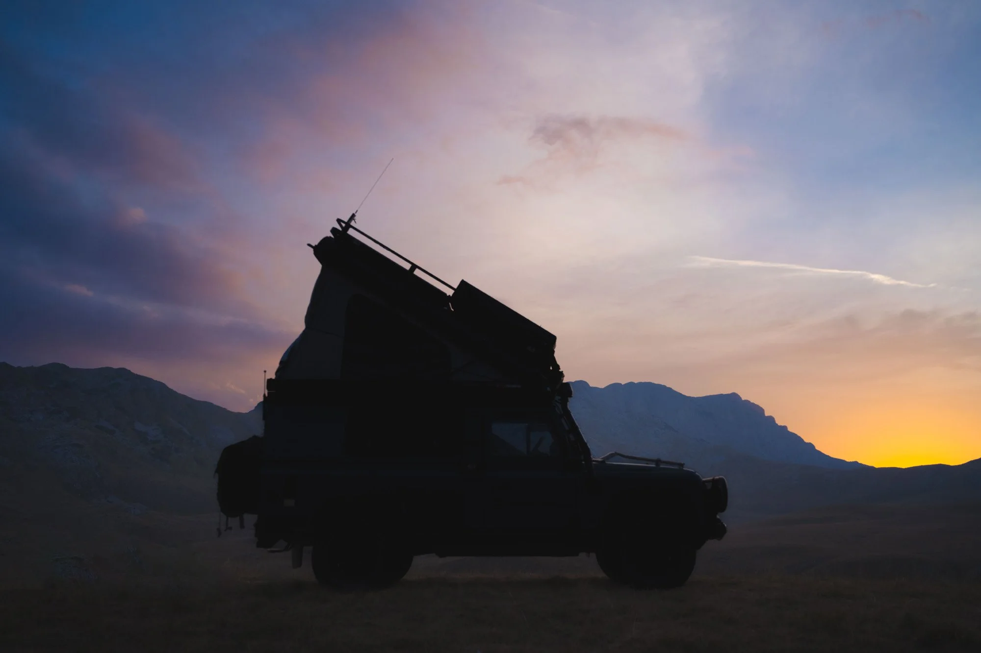 A 1997 Land Rover Defender expedition vehicle silhouetted against a mountain sunset in Montenegro. The simplified graphic shape of the vehicle acts as a strong visual anchor against the complex, atmospheric sky.