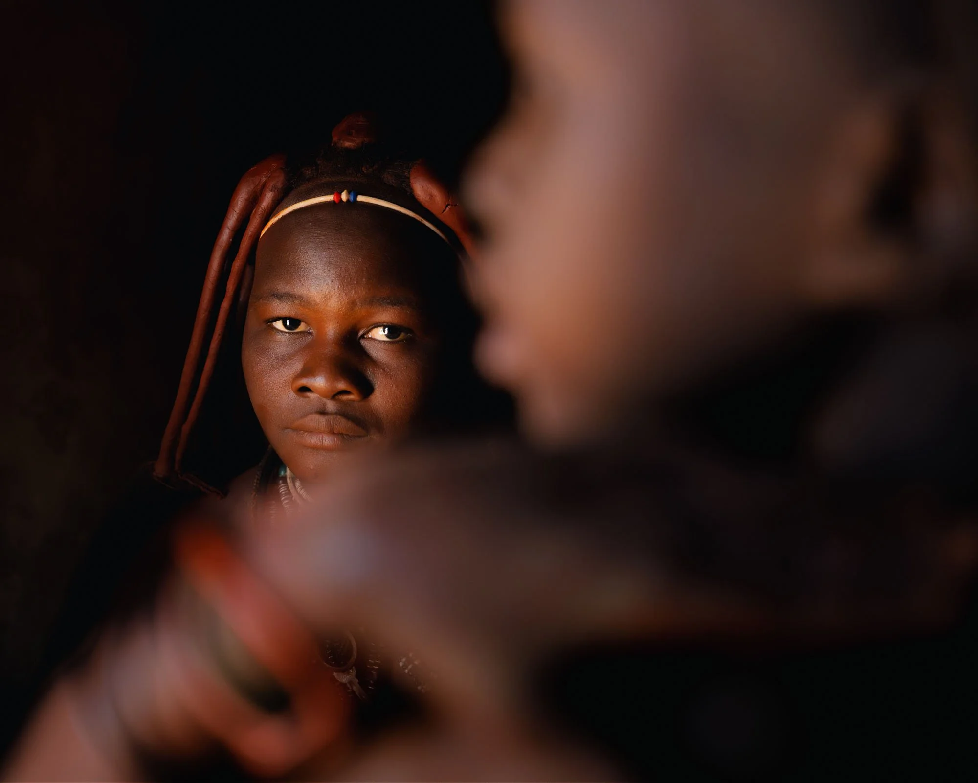 A low-key portrait of a young Himba woman emerging from deep shadow with soft side-light defining her facial expression in Namibia