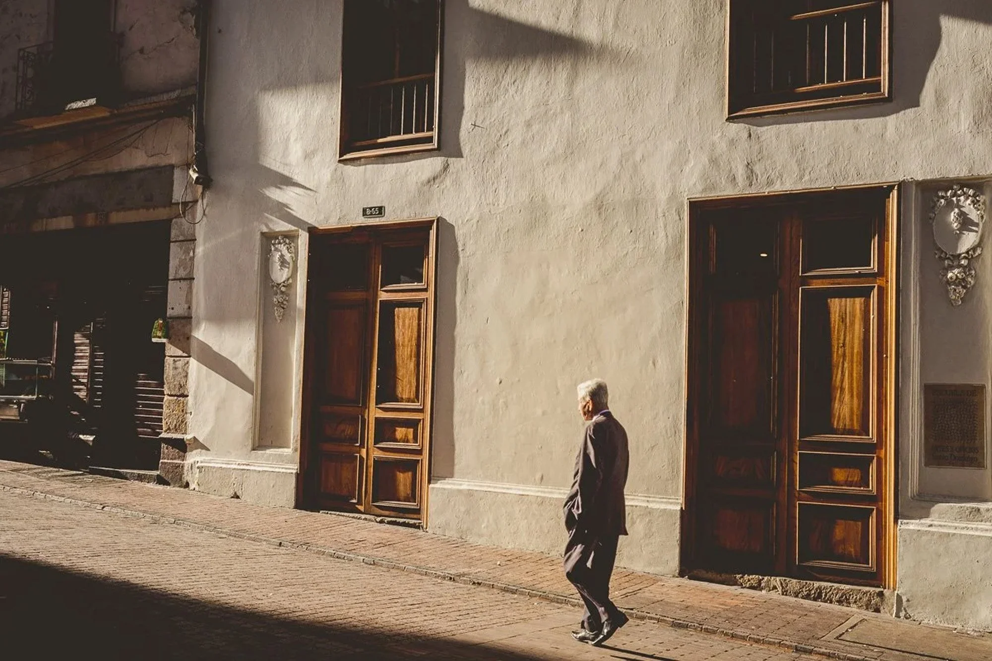 An elderly man dressed in a suit walking along a sunlit sidewalk outside a building with large wooden doors and windows, casting shadows on the wall.