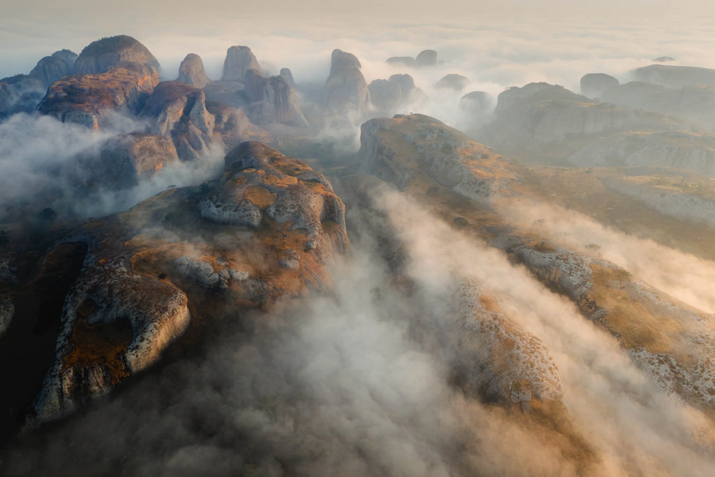 Aerial view of foggy mountain peaks with rugged rocky terrain and sparse vegetation, enveloped in clouds and mist.