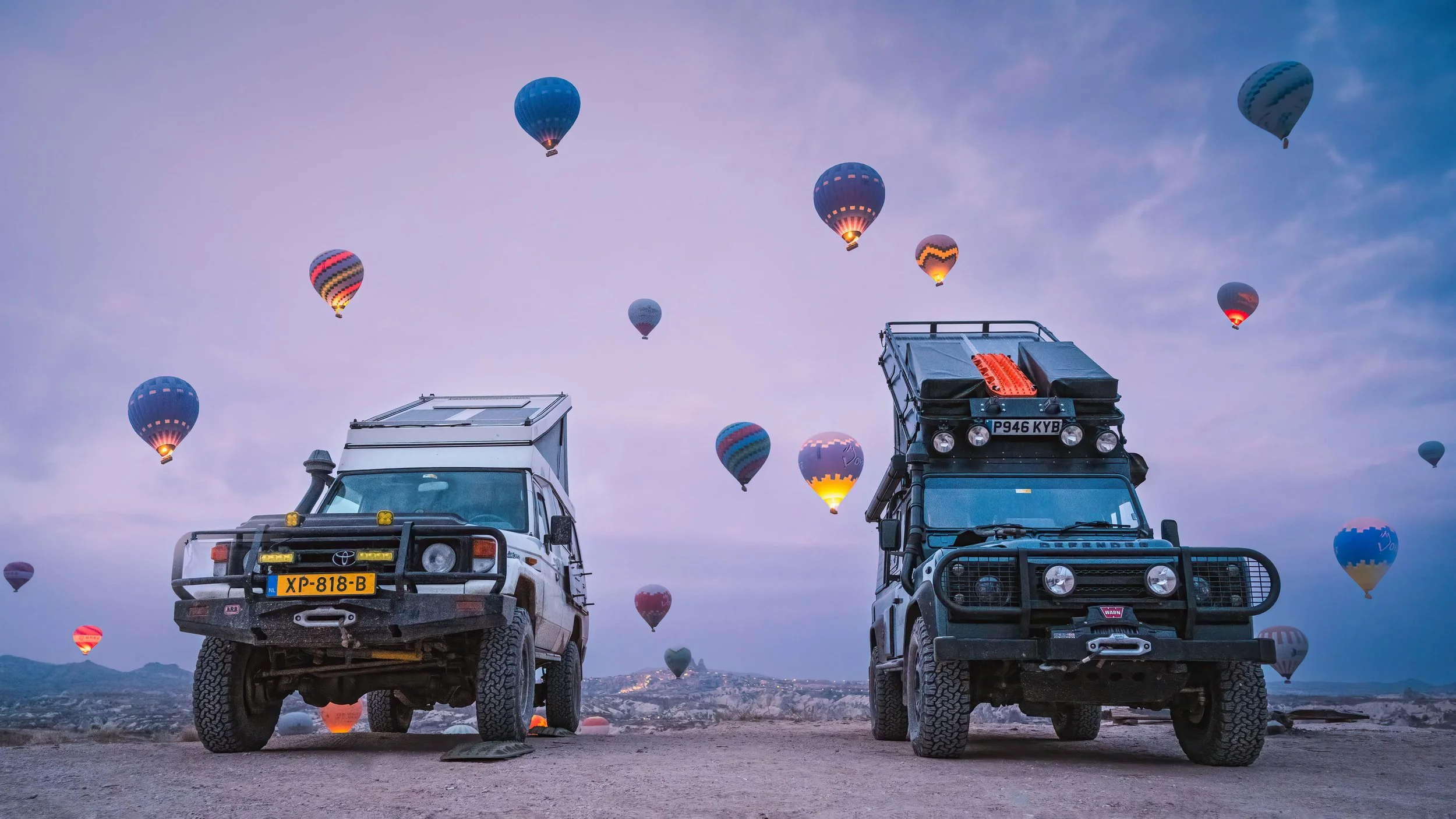 Two off-road vehicles parked on a dirt surface with hot air balloons flying in the sky during dusk.