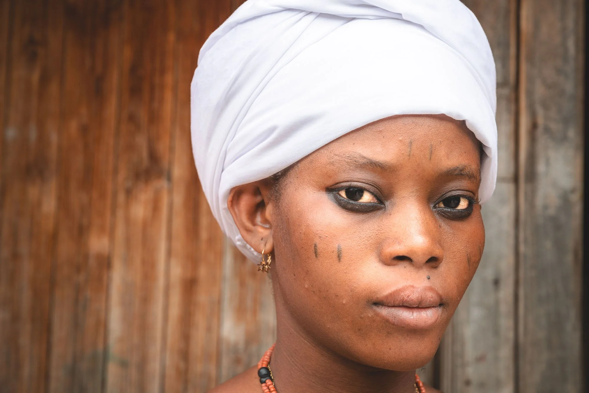 Close-up portrait of a Vodun Queen from Savalou, Benin, with traditional facial scarification marks, a white headwrap, and beaded necklaces.