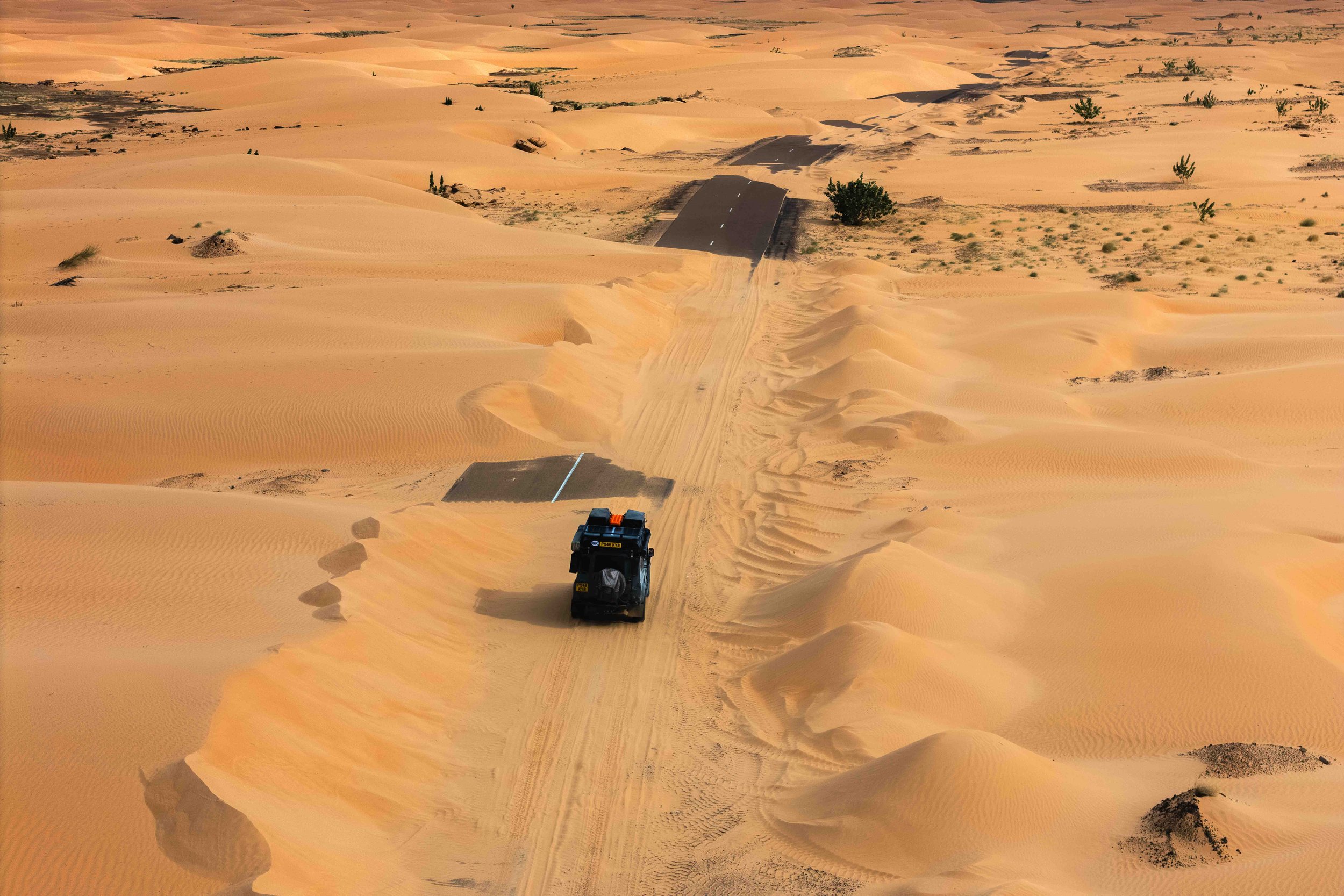 A Land Rover Defender drives down a strip of asphalt being reclaimed by the Sahara Desert dunes in Mauritania. This visual anomaly triggers the seeking system, reminding the observer that the journey is the real art.