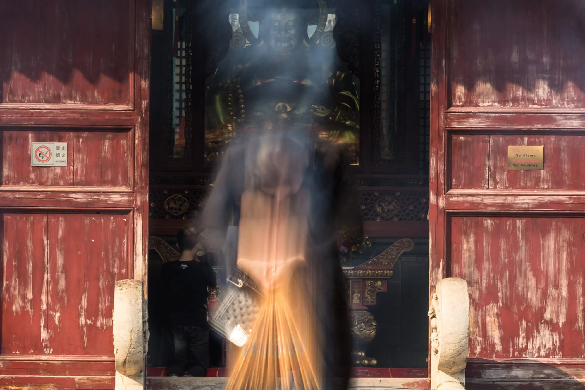 Worshiper moving from long exposure surrounded by heavy, swirling clouds of burning incense smoke illuminated by morning sunlight at Longhua Temple in Shanghai