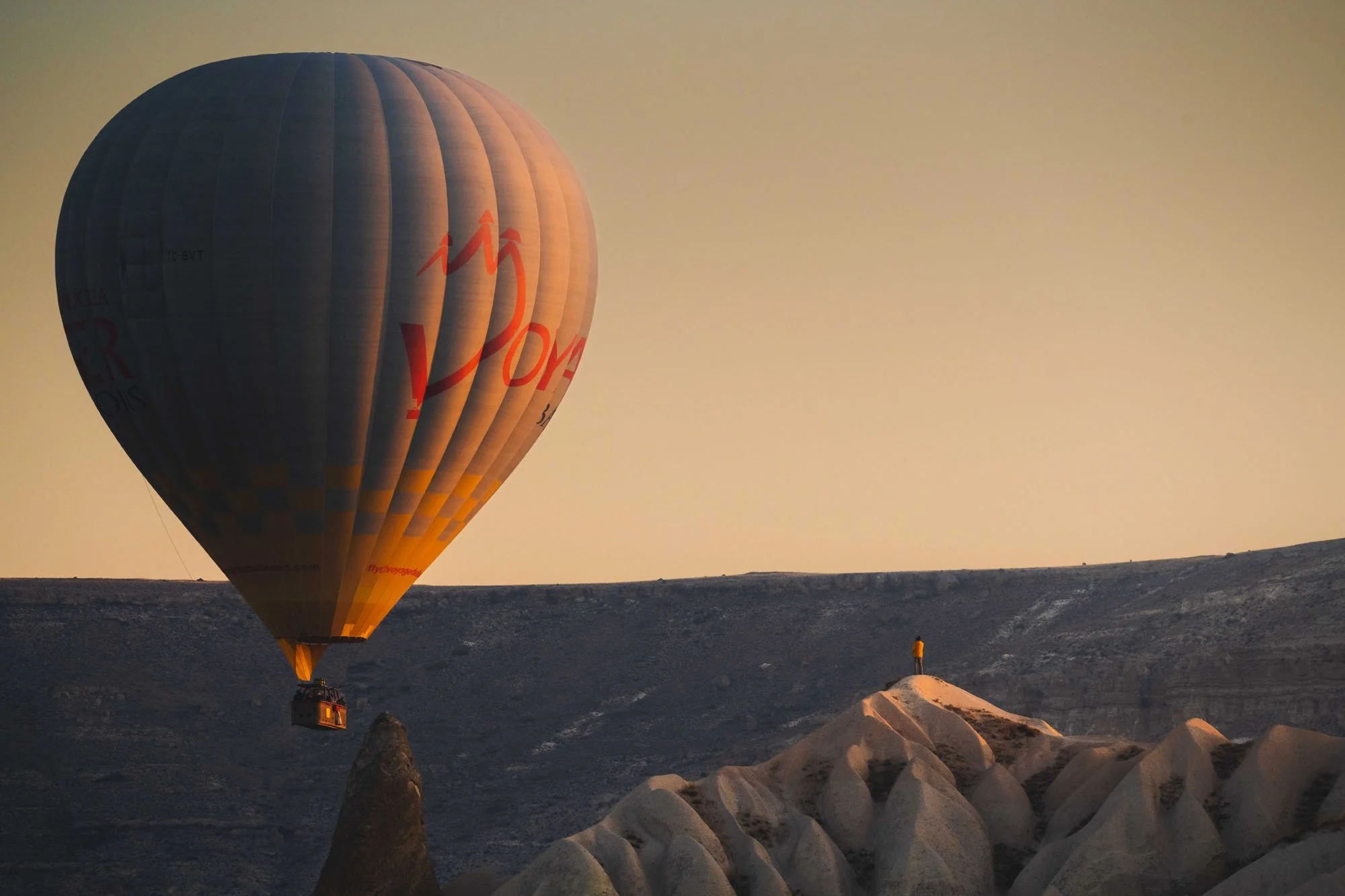 A massive hot air balloon in the foreground with a microscopic human figure standing on a distant ridge in Cappadocia, using scale as a biological anchor to prove environmental immensity