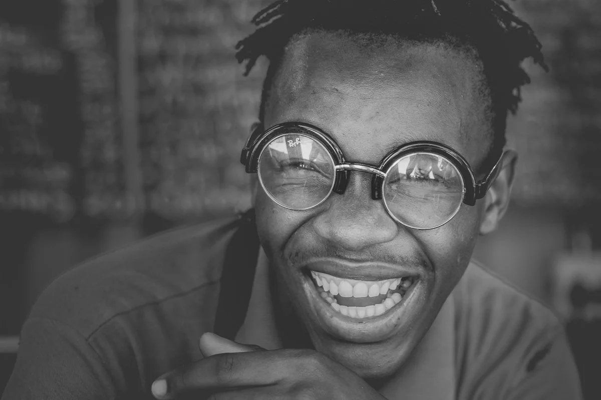 Close-up black and white documentary portrait of a young man laughing joyfully with round glasses in Langa Township, South Africa