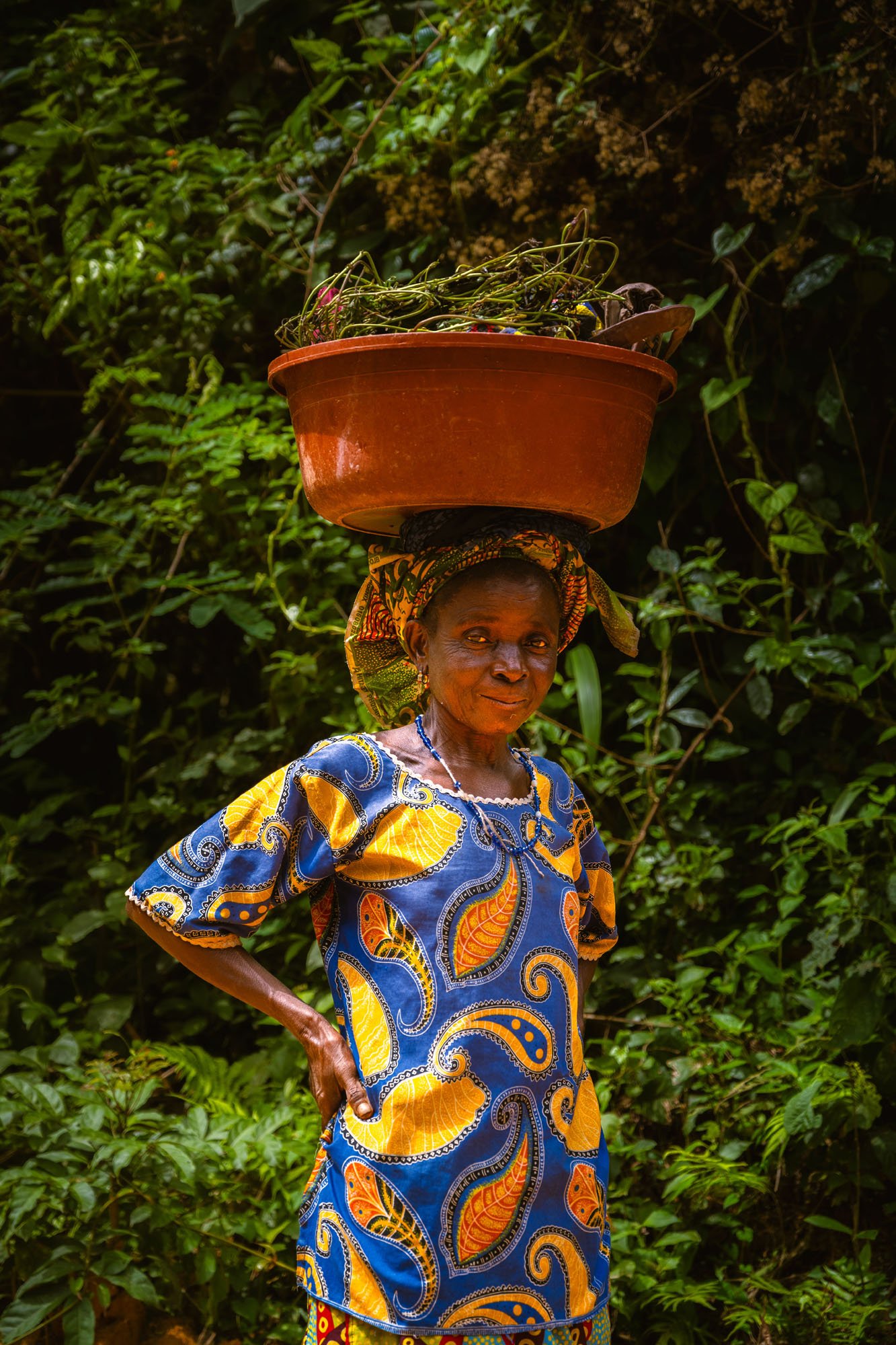 A confident portrait of a woman in vibrant West African textiles balancing a basin on her head. The direct eye contact and authentic environment serve as a 'schema violation' against polished travel clichés.