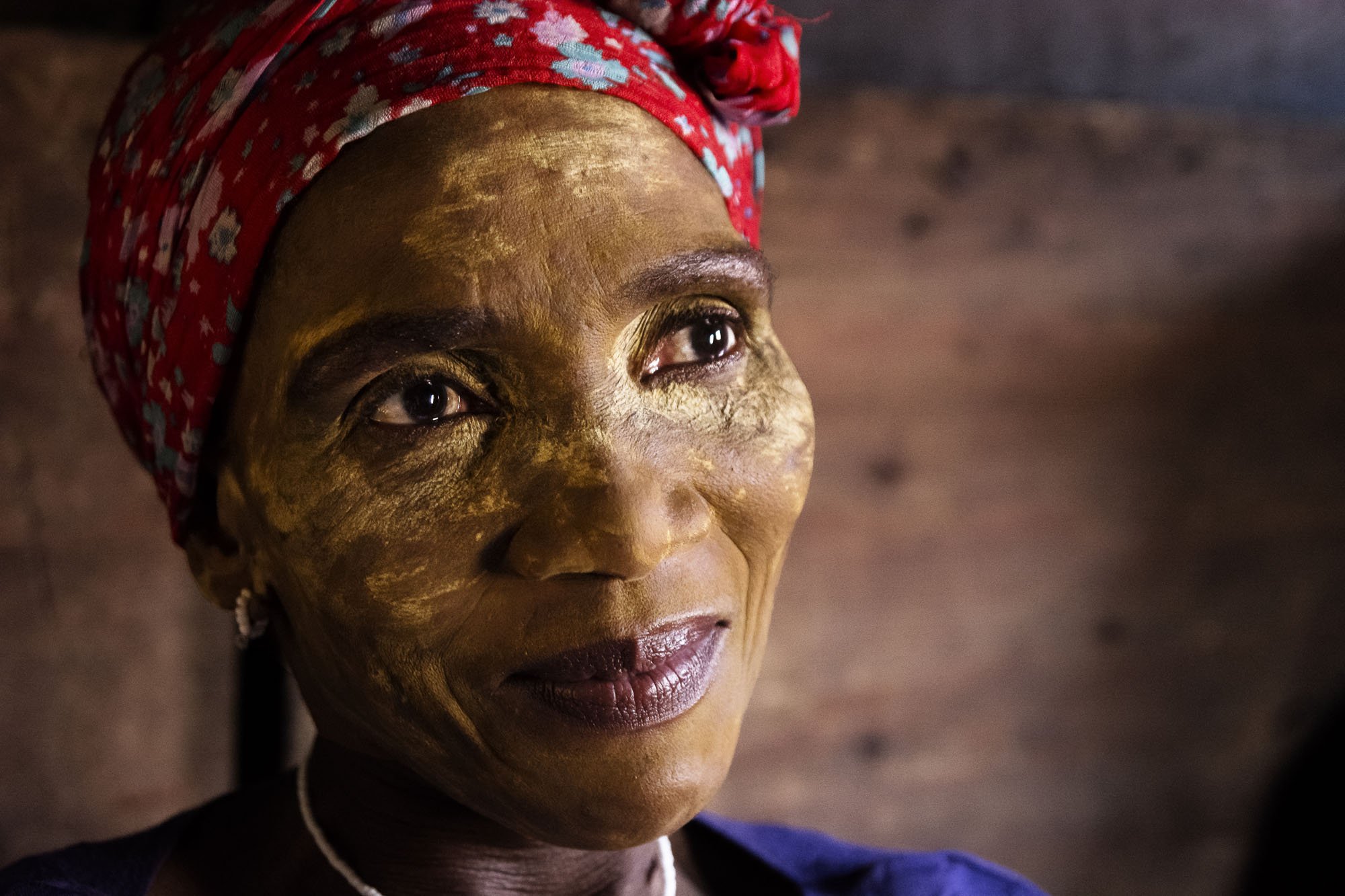An intimate close-up documentary portrait of an older South African woman with traditional yellow clay paste on her face, illuminated by warm directional light inside a Langa shebeen