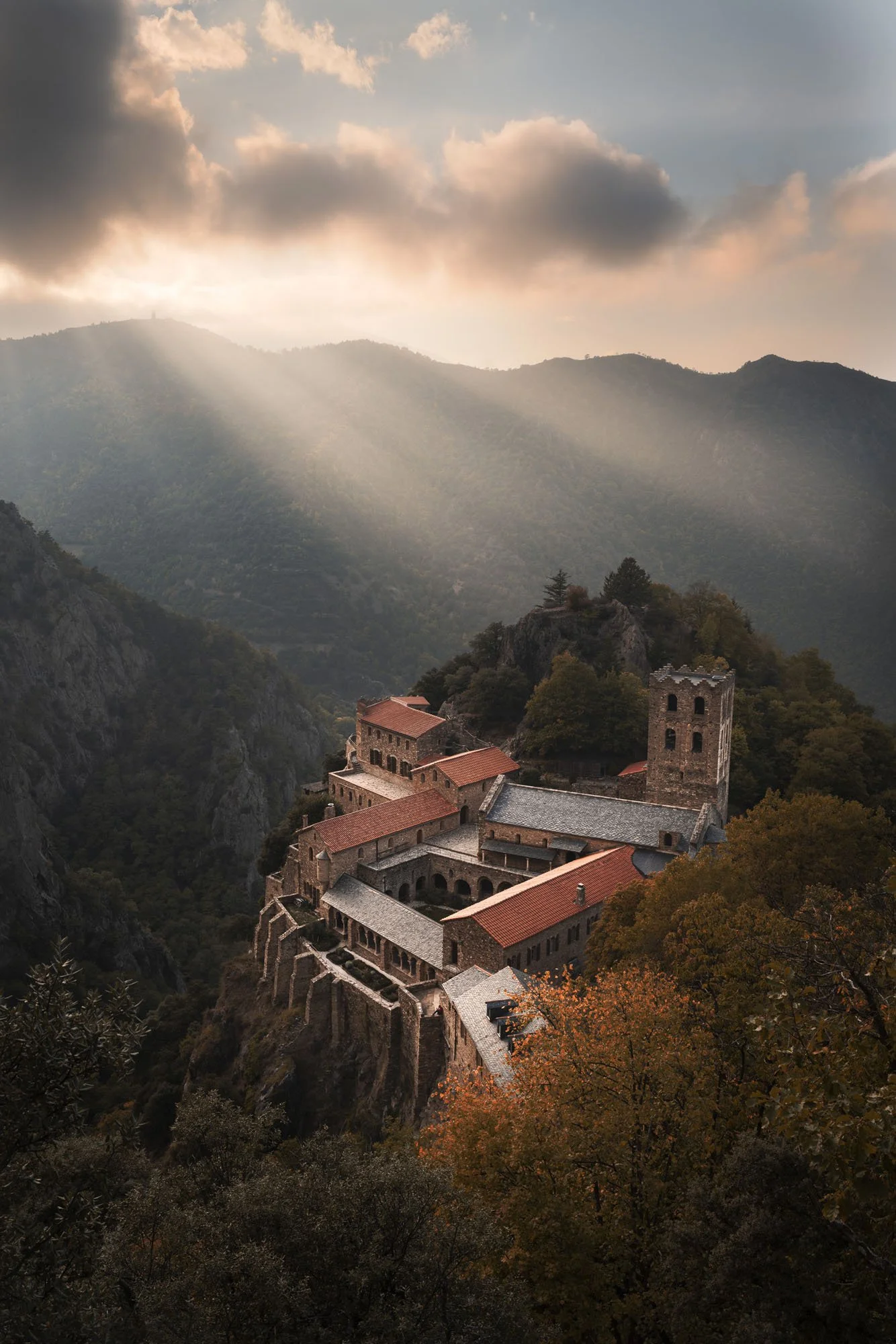Dramatic shafts of celestial sunlight breaking through dark clouds onto an ancient stone monastery perched on a mountain cliff in the Pyrenees