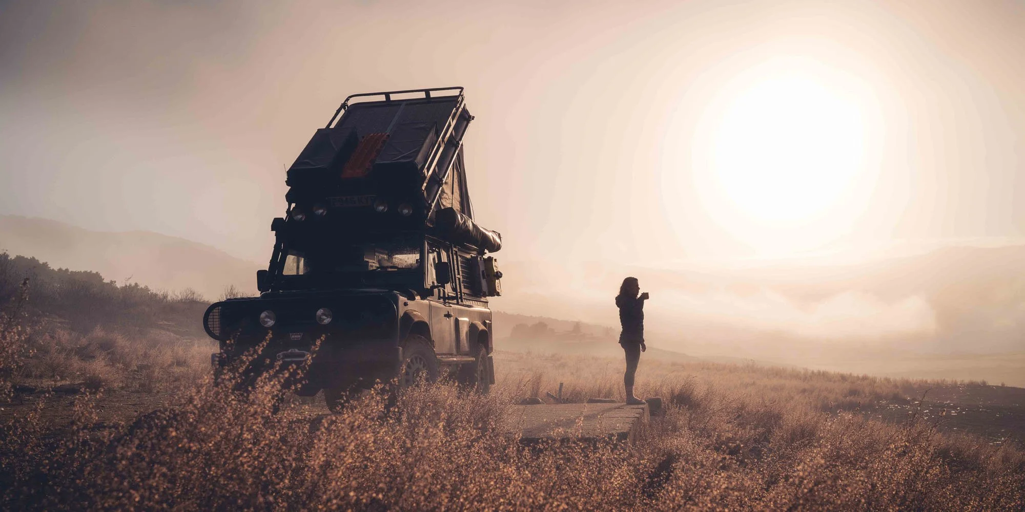 A Land Rover Defender parked at a misty sunrise mountain camp, with a person standing in the tall grass. This image represents the 'horizon'—the world that doesn't care if you take the photo, only that you are there.