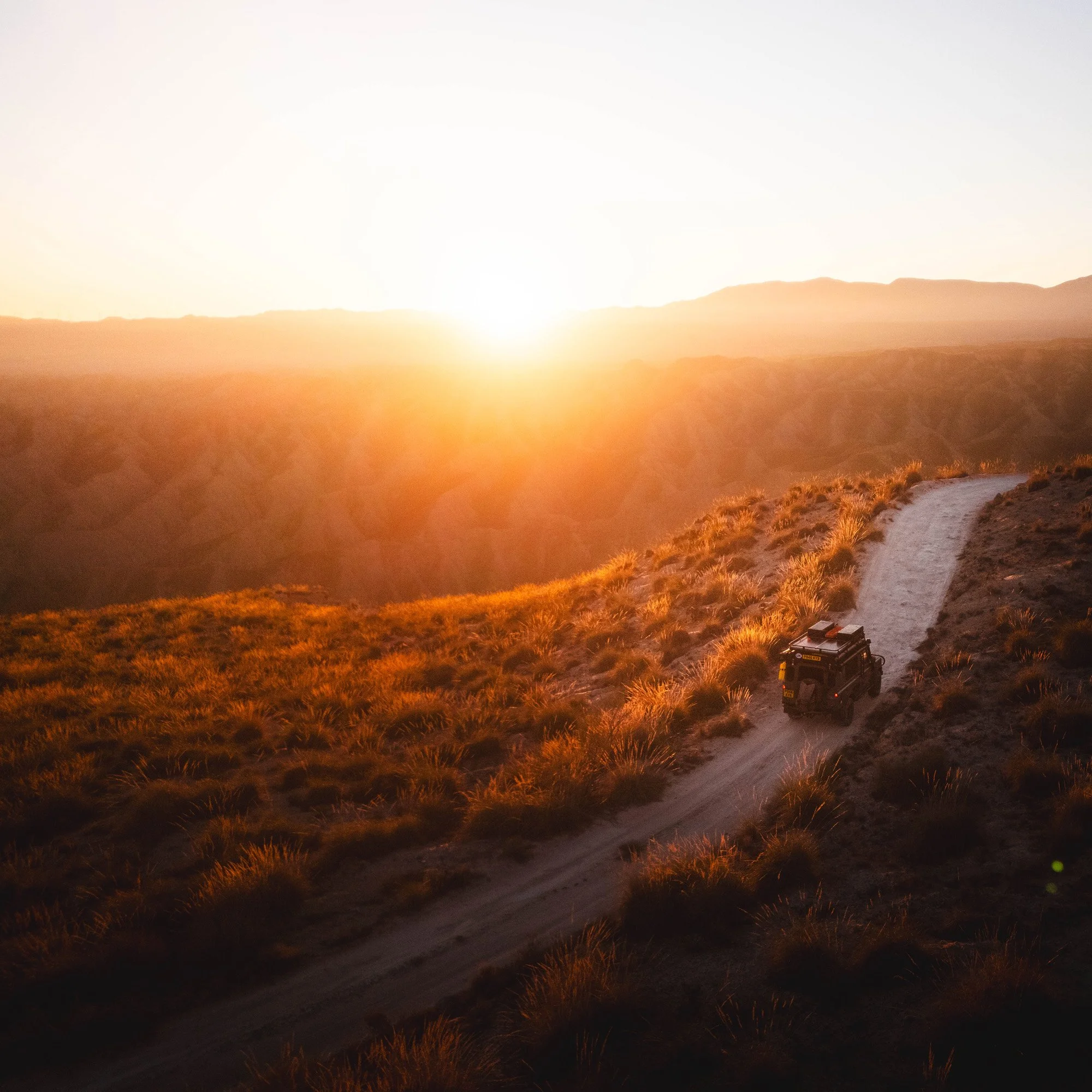 A Land Rover Defender drives along a high-contrast dirt ridge at sunset. The winding track acts as a visual breadcrumb (leading line) that forces the viewer to exert cognitive labor to follow the vehicle's path toward the horizon.