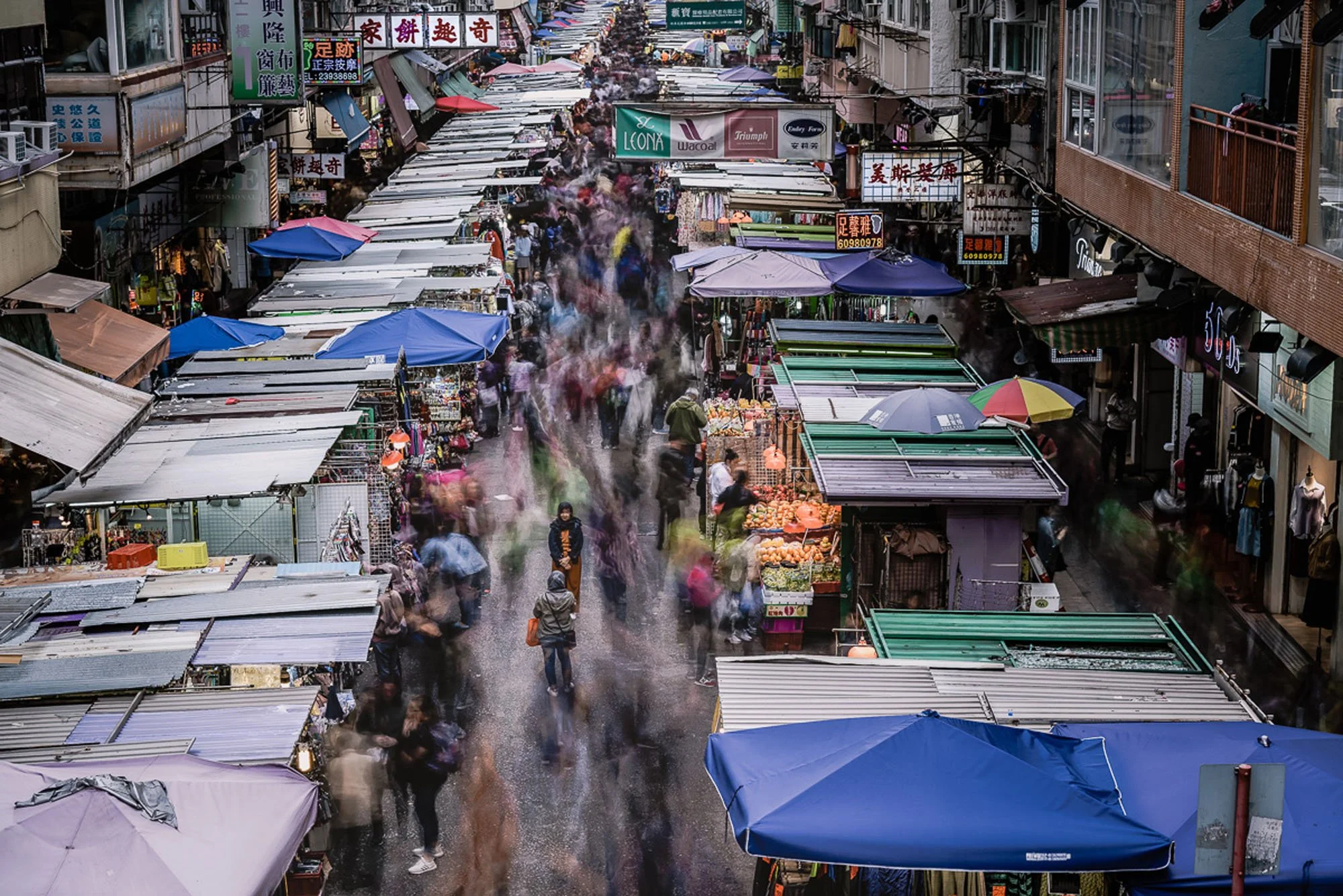 An overhead view of a dense, bustling street market in Hong Kong with motion-blurred pedestrians surrounding a single still figure
