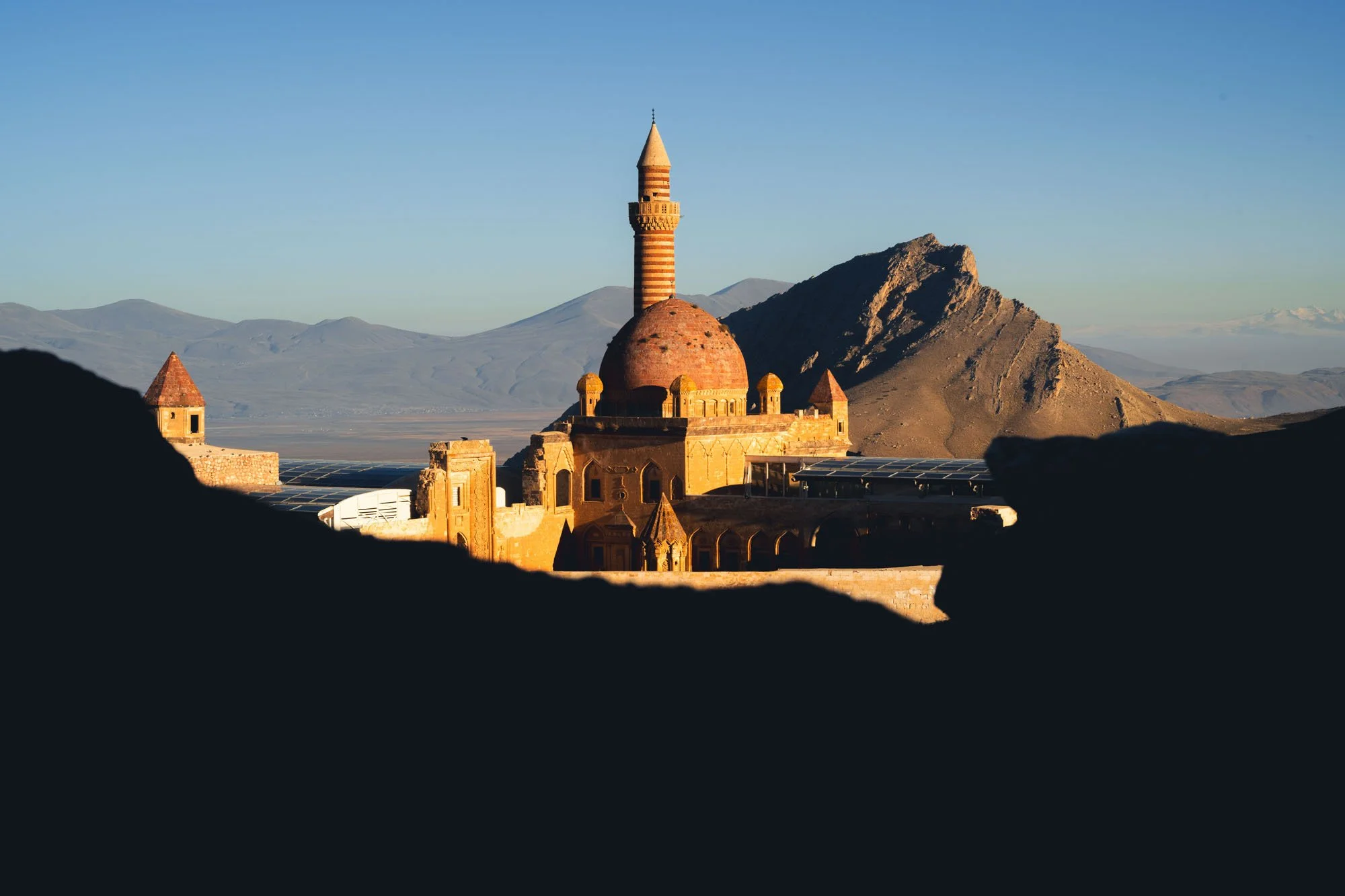 The sunlit dome and minaret of the historic Ishak Pasha Palace in Eastern Türkiye framed dramatically by the dark, cavernous silhouette of surrounding rocks