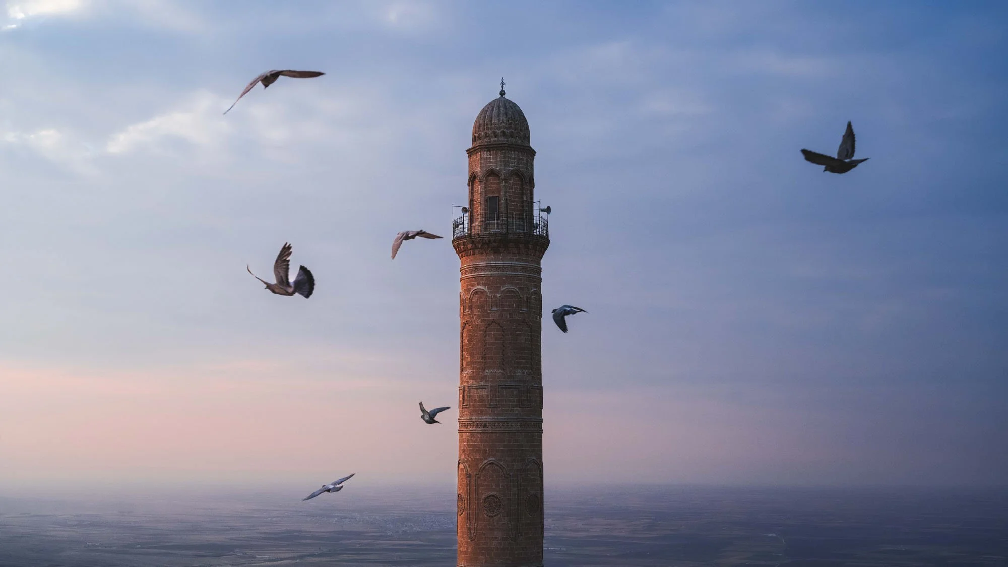 A tall stone minaret in Mardin, Türkiye, with pigeons in flight against a blue dusk sky, showing a visual signal that refuses to tell a traditional story.