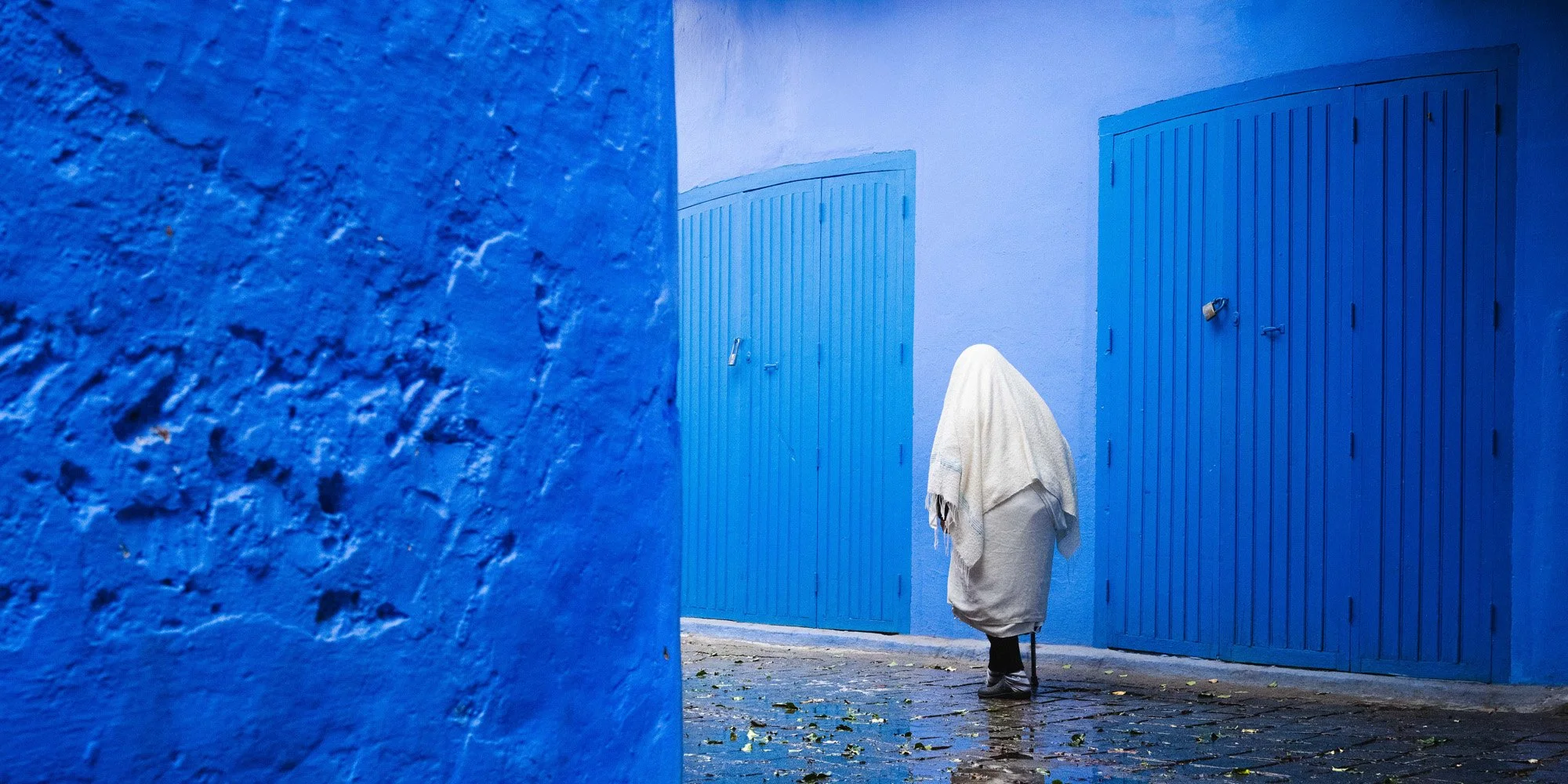 A figure draped in white walks past the iconic vibrant blue doors and walls of Chefchaouen, Morocco. The extreme color contrast and partially obscured subject create visual ambiguity, triggering a dopaminergic 'hunt for meaning' in the brain.
